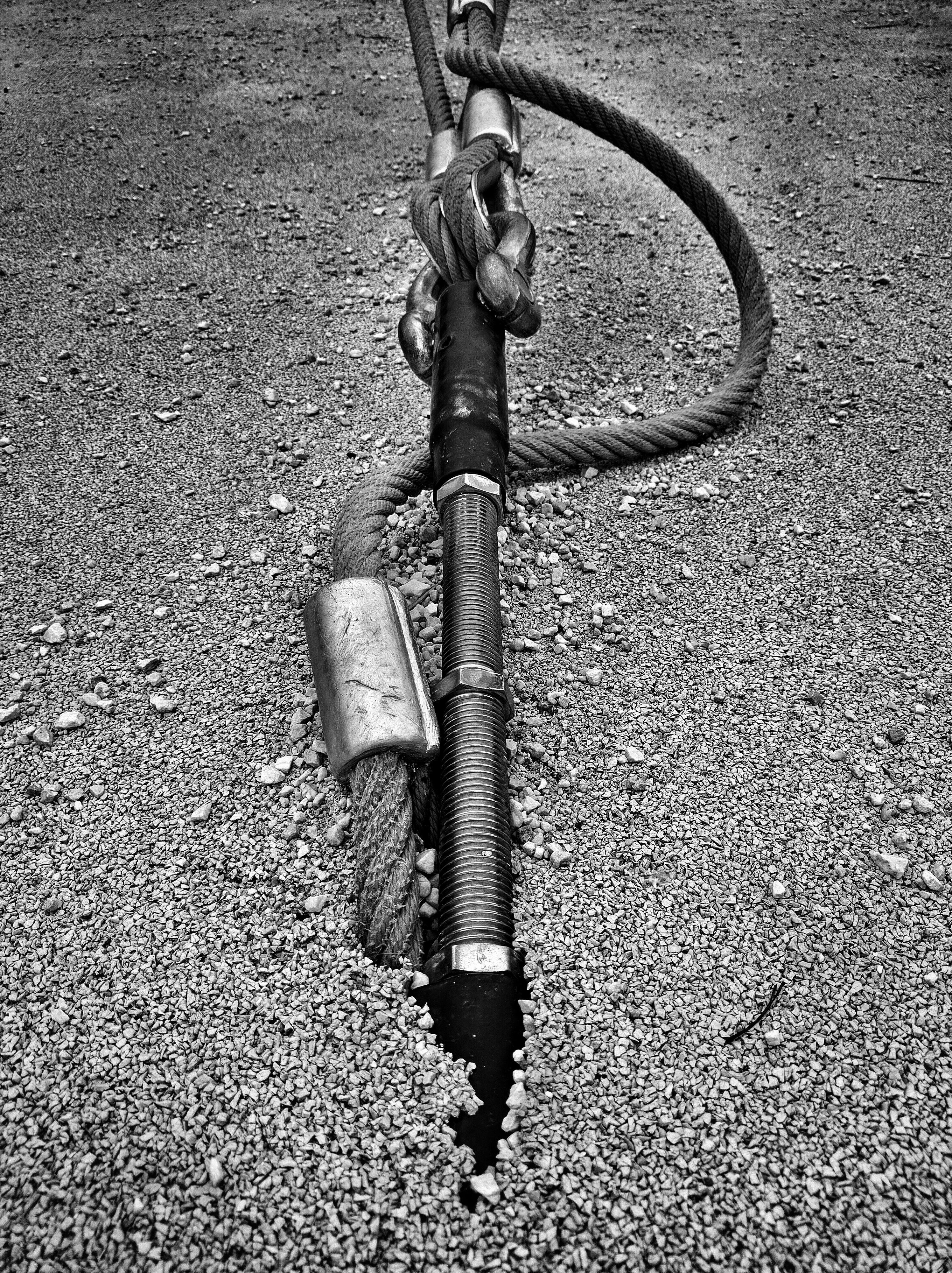 The image shows a close-up of a thick, coiled metal cable or rope lying on a gravel surface. The cable is wrapped around a black rubber hose with metal fittings and clamps. The gravel looks loose with some small pebbles scattered around. The setup suggests it's part of some industrial or construction setup.
