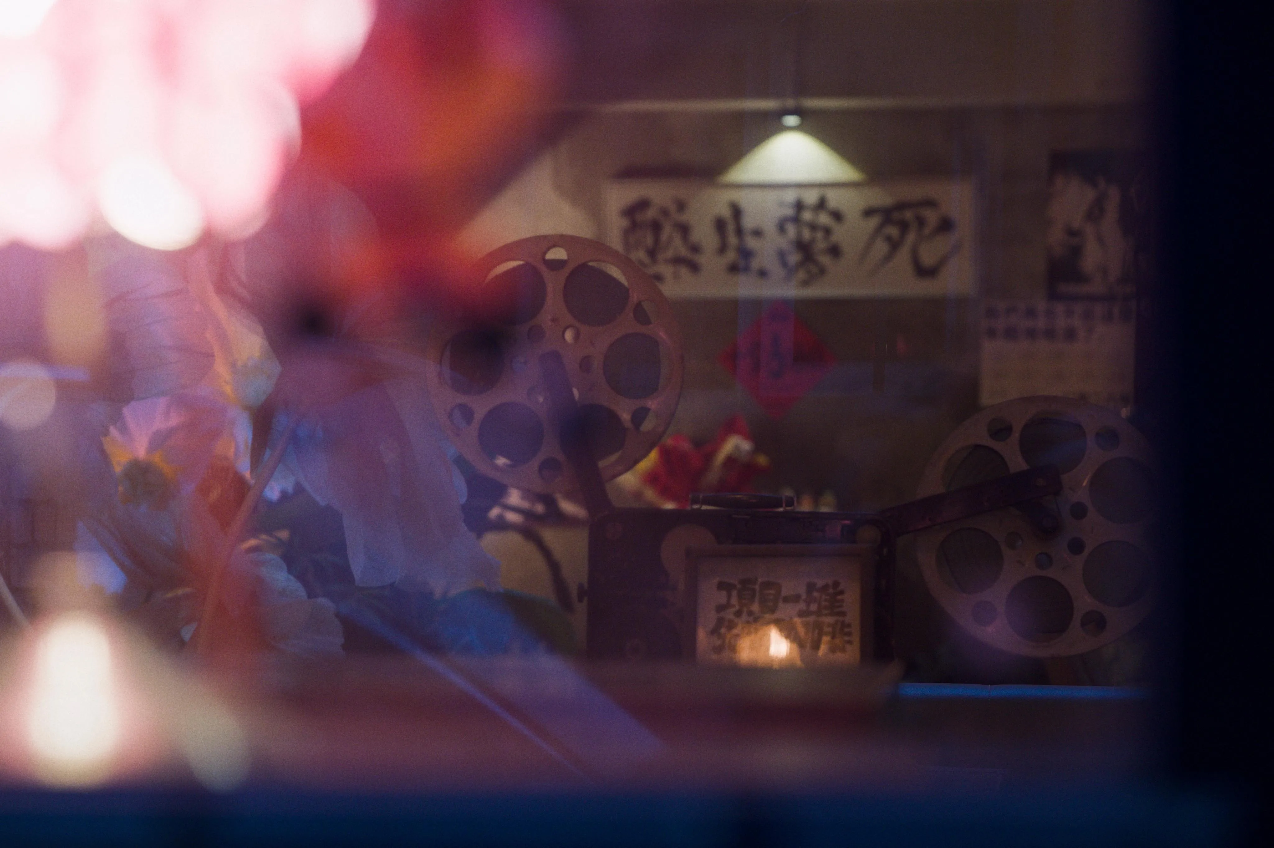 Photograph of a vintage film projector taken from outside a window with reflections, tinting the image with various colors. Flowers with large blue petals are disposed besides the film projector, but cannot be seen clearly because of the foreground reflections.
