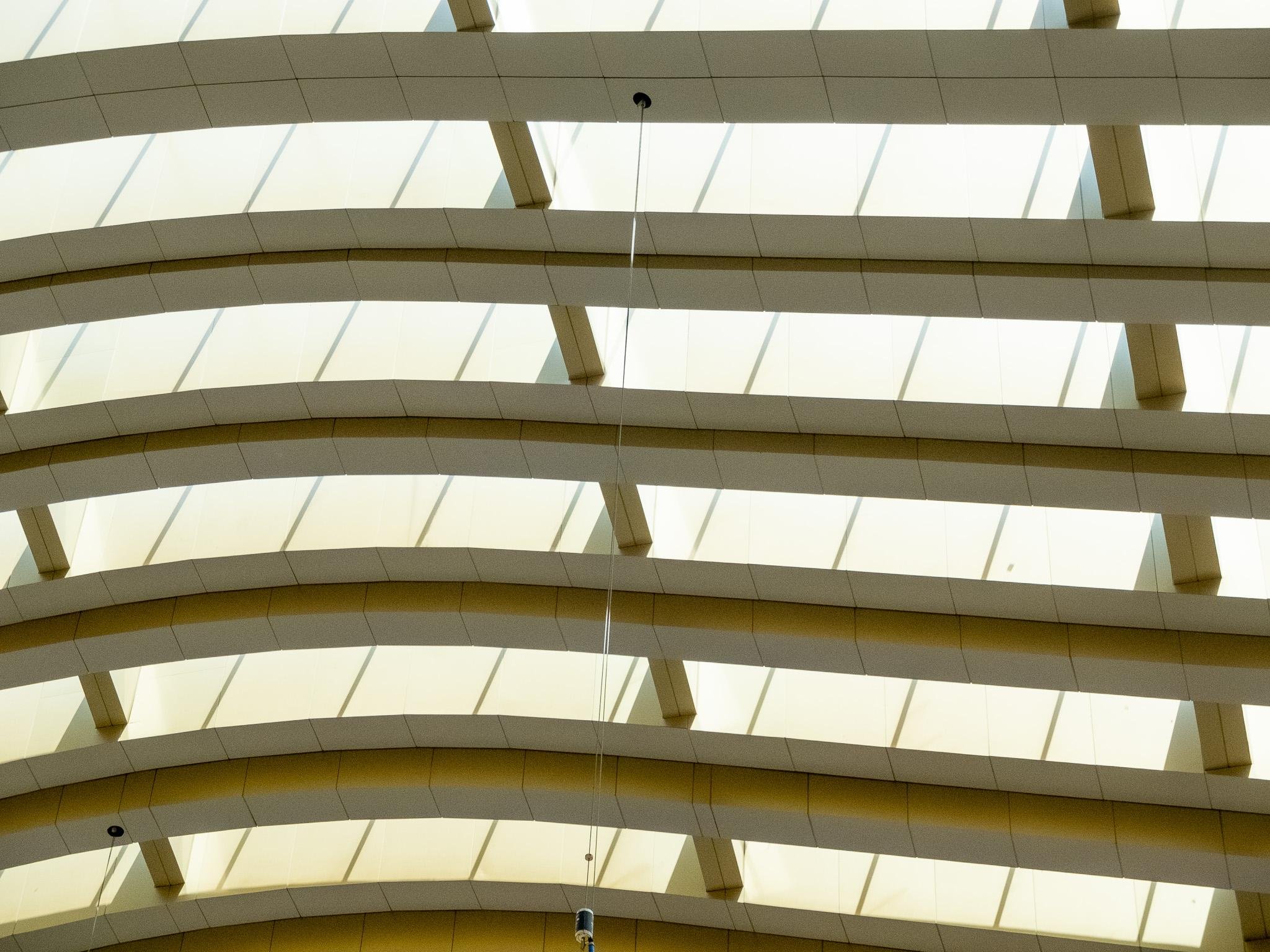 The image shows the ceiling of a building with a modern, architectural design. It consists of multiple glass panels set in a grid-like pattern, allowing light to pass through. The structure has curved beams or arches, giving it an aesthetic and contemporary appearance.
