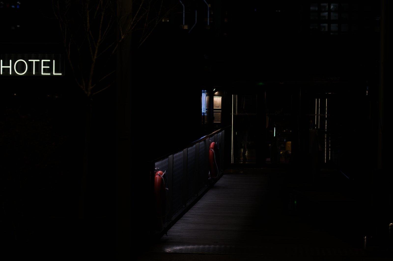 Photo nocturne très sombre mais avec une enseigne «&nbsp;HOTEL&nbsp;» lumineuse et sur la droite, une passerelle faiblement éclairée, avec des barrières de sûreté où sont accrochées deux bouées de sauvetage rouge, menant à une porte avec les trait de lumières or verticales de chaque côté