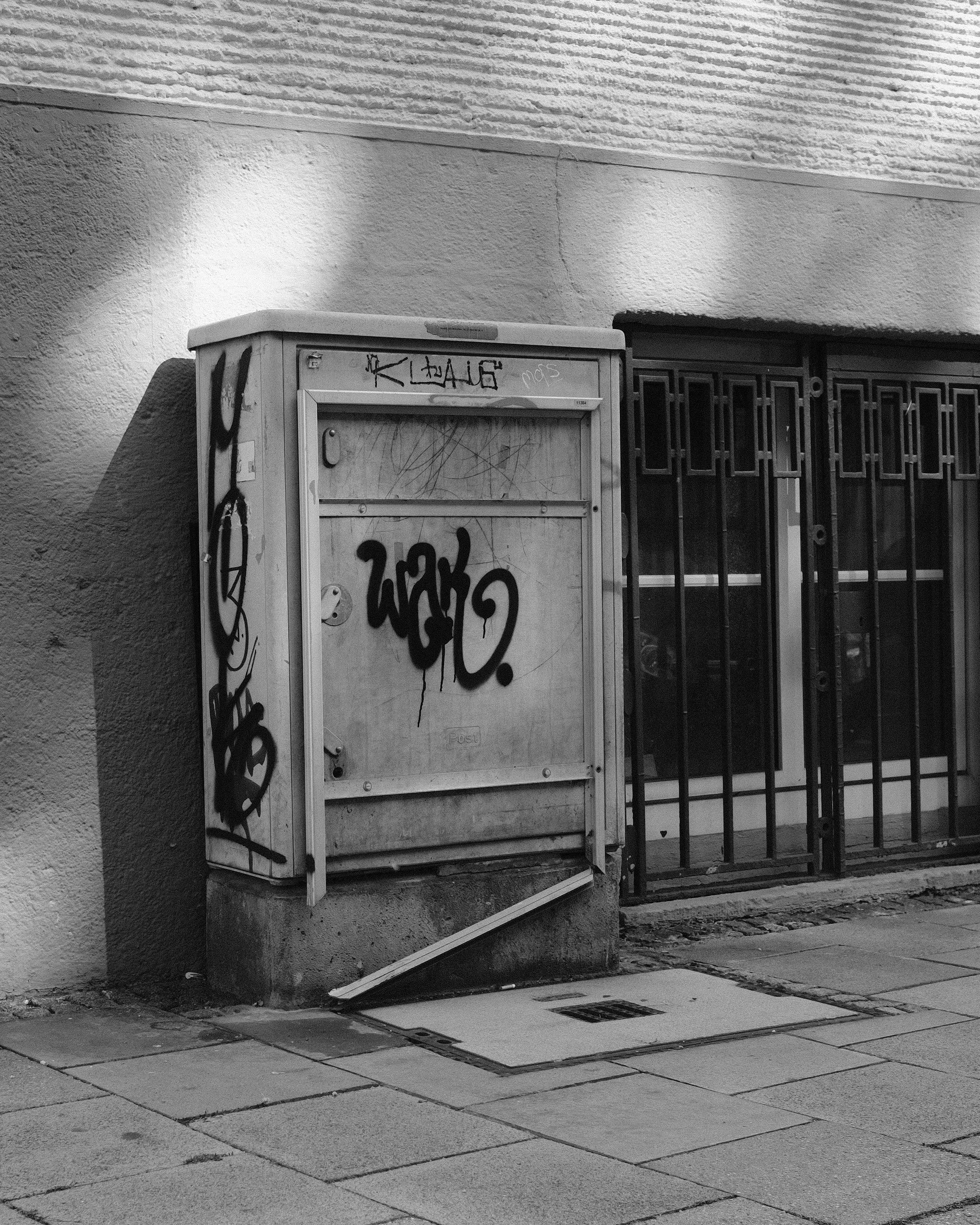 Graffiti-covered and damaged utility box on a sidewalk against a wall. Next to it is a barred window. Patches of sunlight and shadow across the wall and pavement.
Monochrome black/white image.g