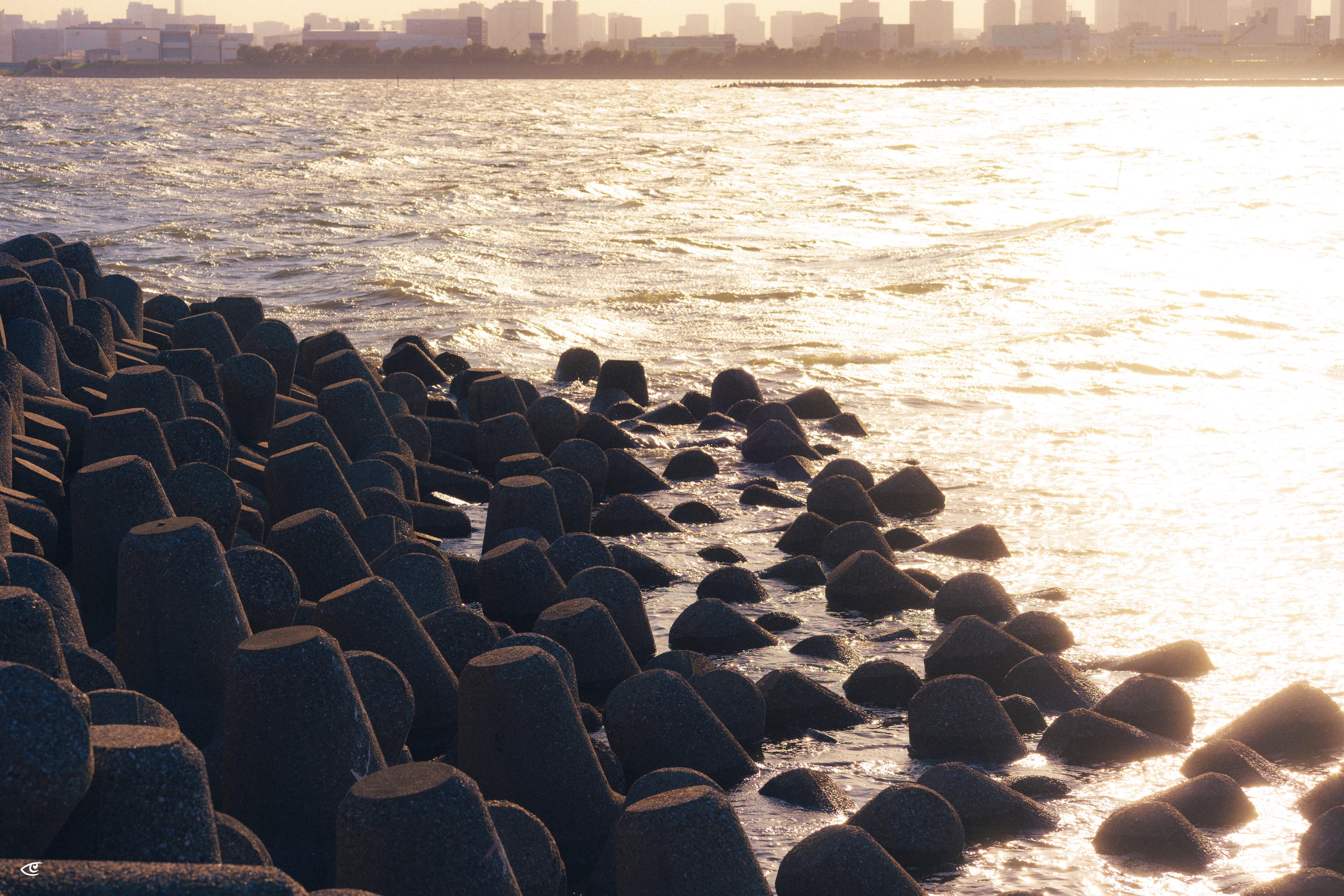 Concrete wave-breaker blocks along a shoreline with small waves washing between them, bright sun glare on the water to the right, and a distant city skyline on the horizon.