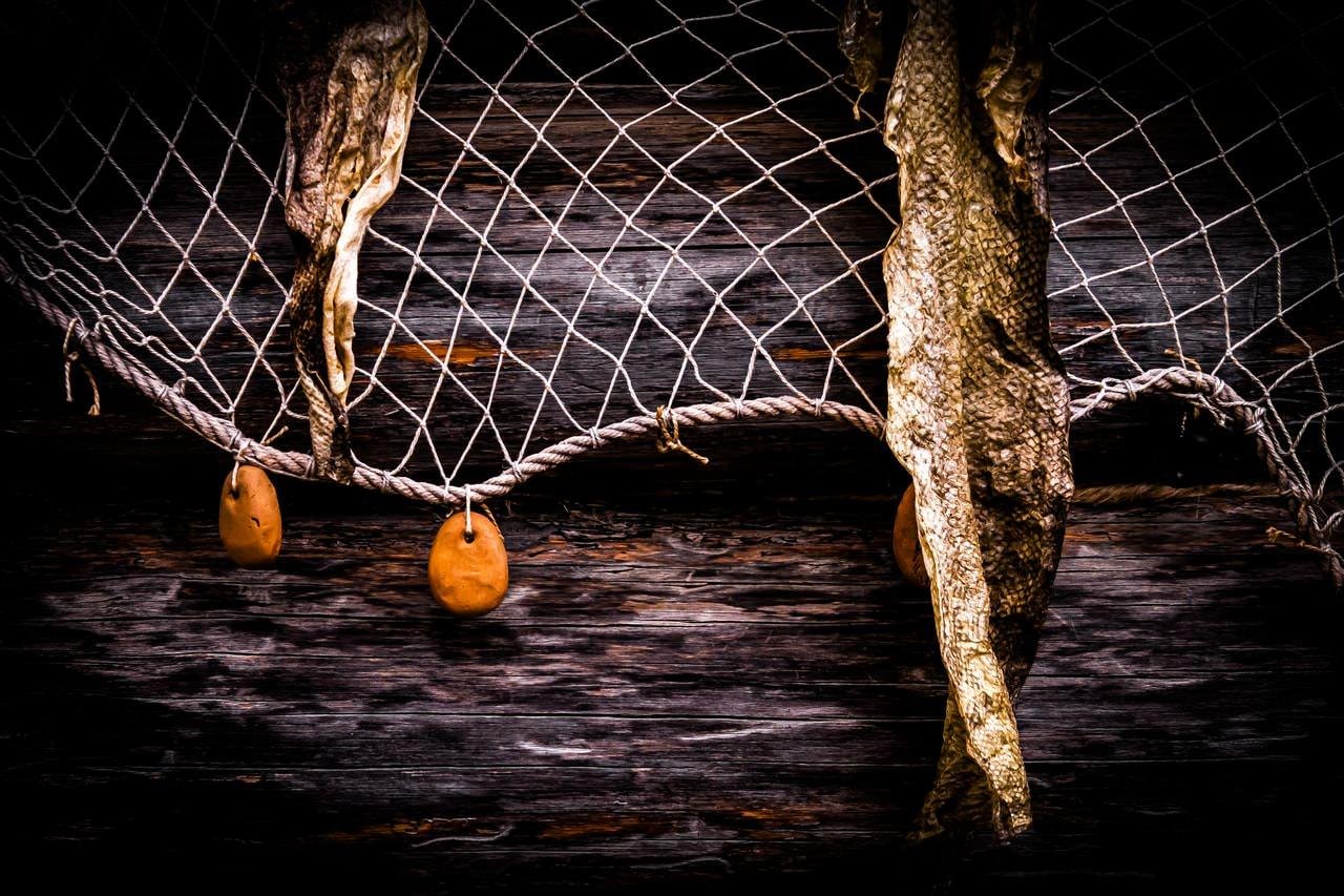 Fishing net with dried fish hanging, featuring coarse rope and round orange floats. Background consists of dark wooden planks with visible grain and texture.