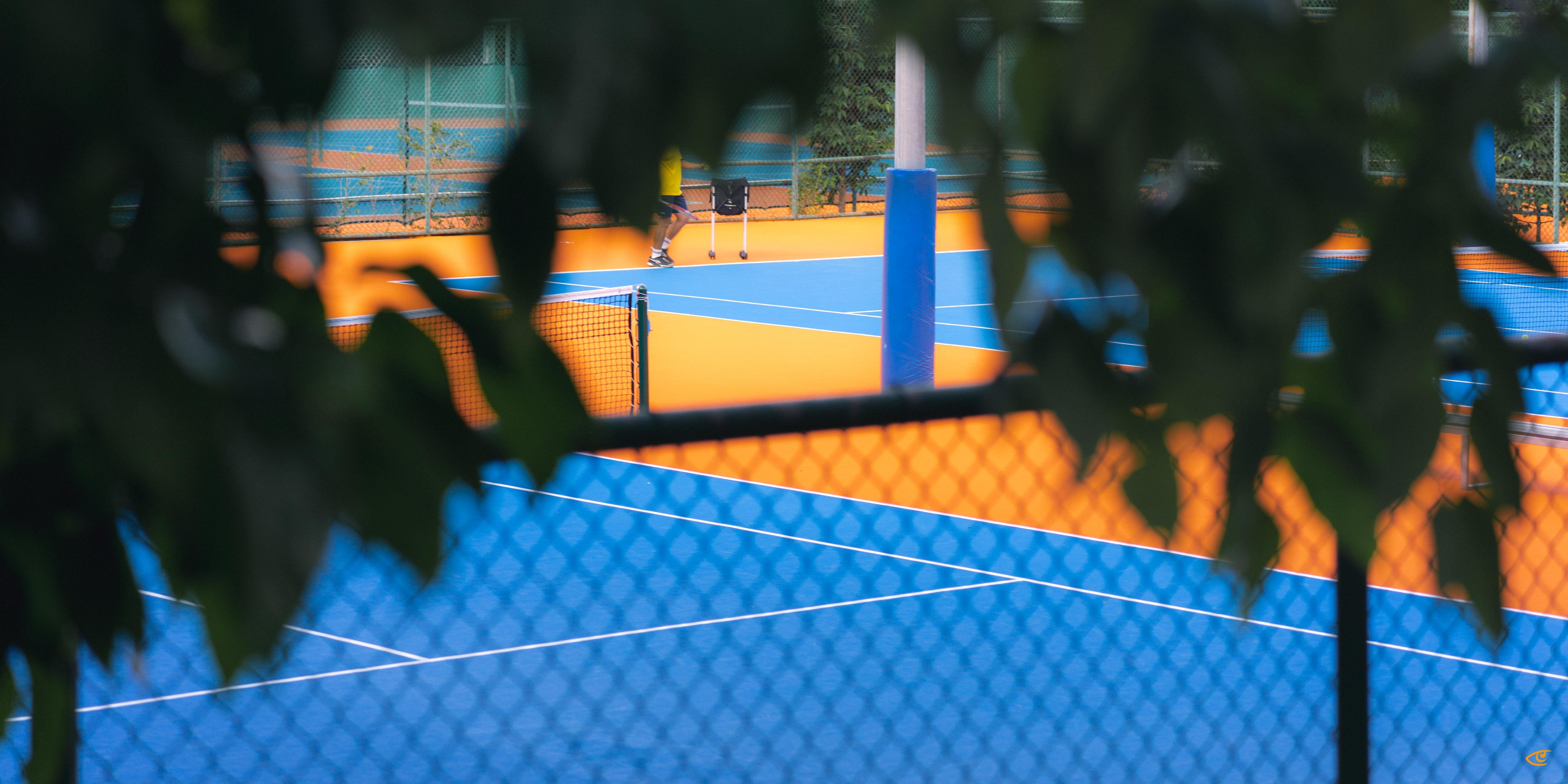 Blue and orange tennis courts viewed through a chain-link fence and out-of-focus tree leaves in the foreground, with a small figure near the far baseline and court net and posts visible.
