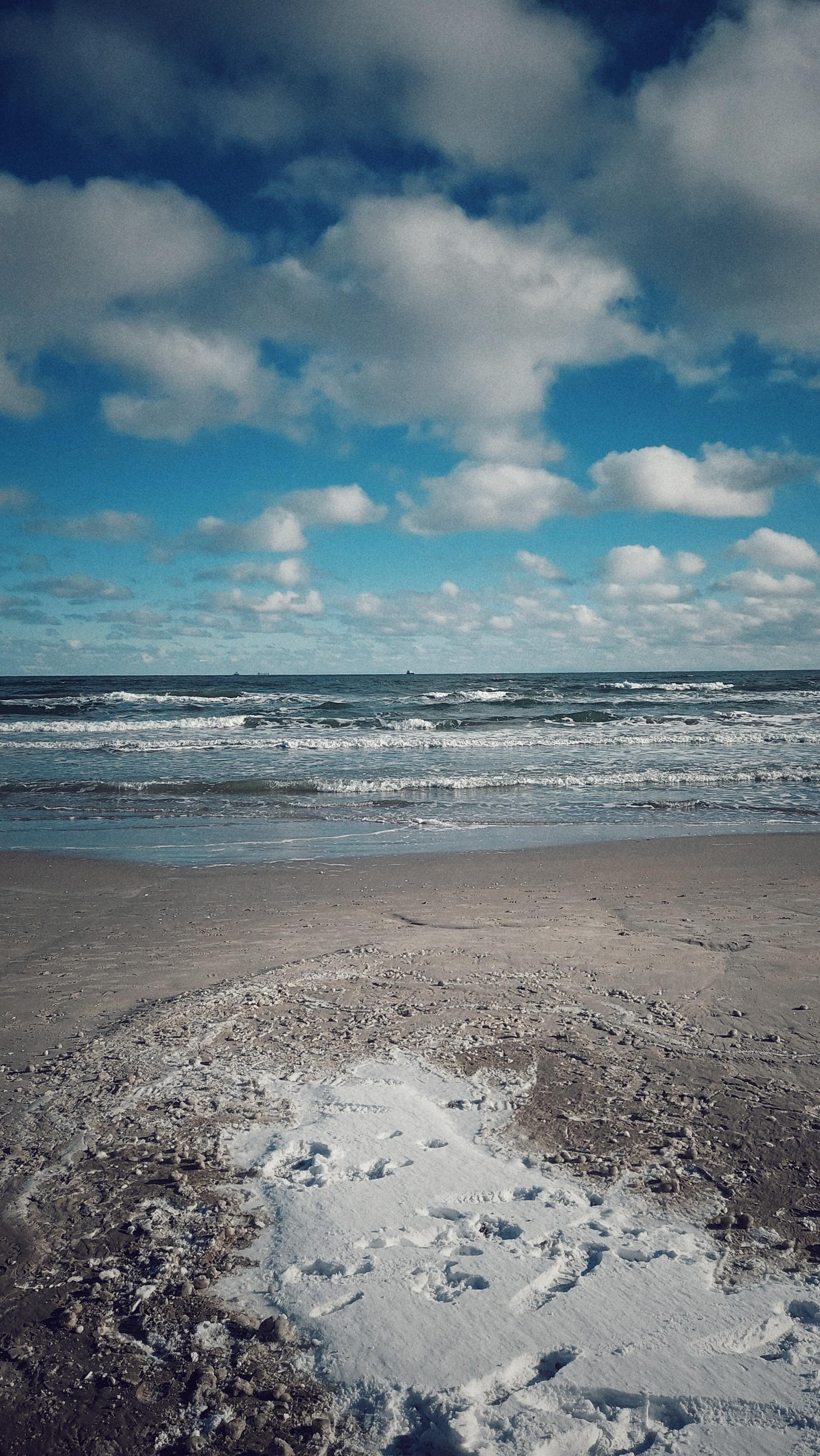 The image shows a beach scene with waves in the ocean and a partly cloudy sky. There is snow or frost visible on the sandy beach, with footprints scattered across it. The sky is blue with fluffy clouds, creating a contrast with the wintry scene on the beach.