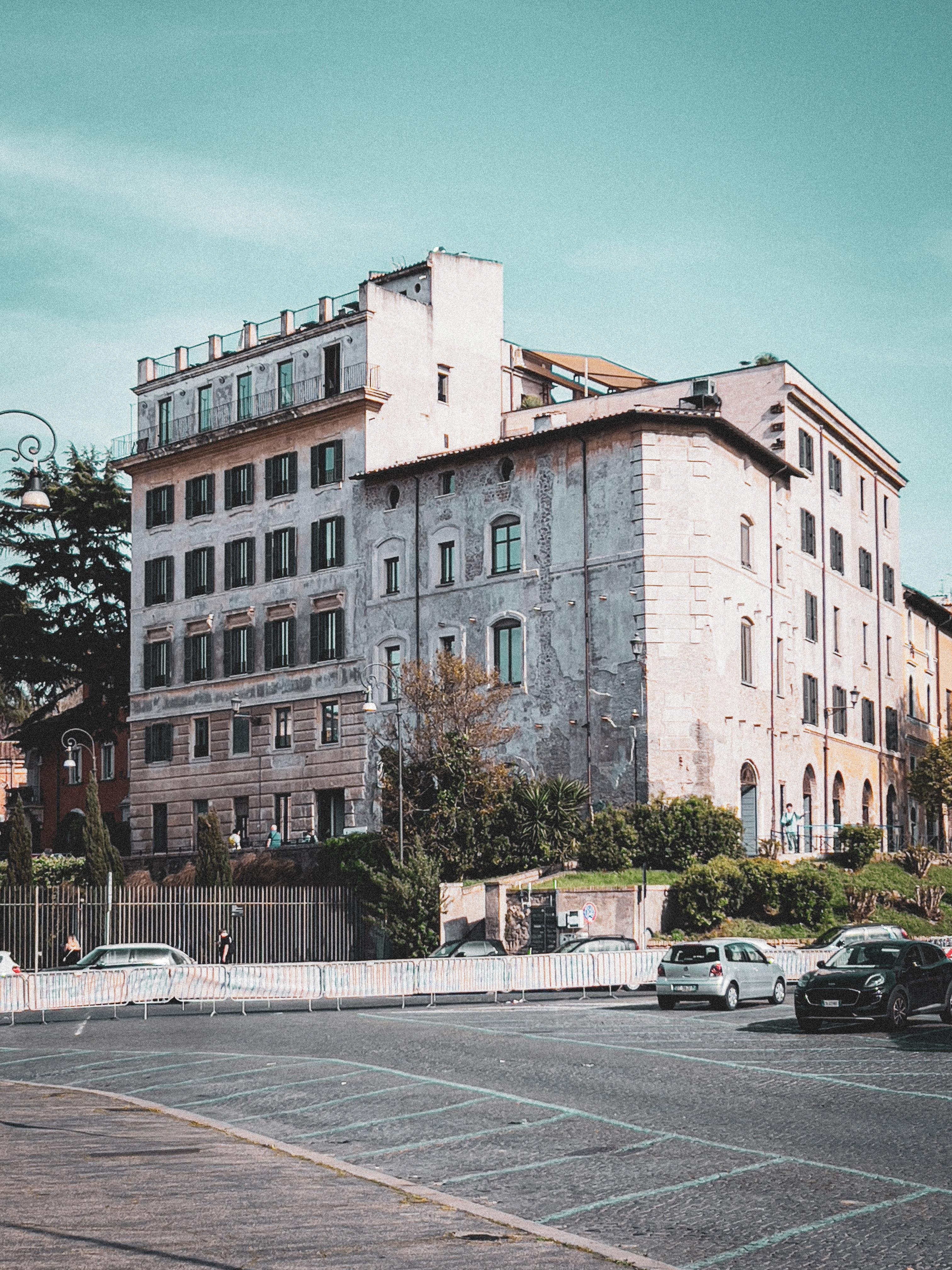 Edificio dall'aspetto storico con finestre ad arco e una facciata usurata, tipica dell'architettura europea più antica. In primo piano, c'è una strada con auto parcheggiate e barriere di metallo. La scena è ambientata in un'area urbana con un po' di verde attorno all'edificio. Il cielo è limpido, suggerendo una giornata soleggiata.