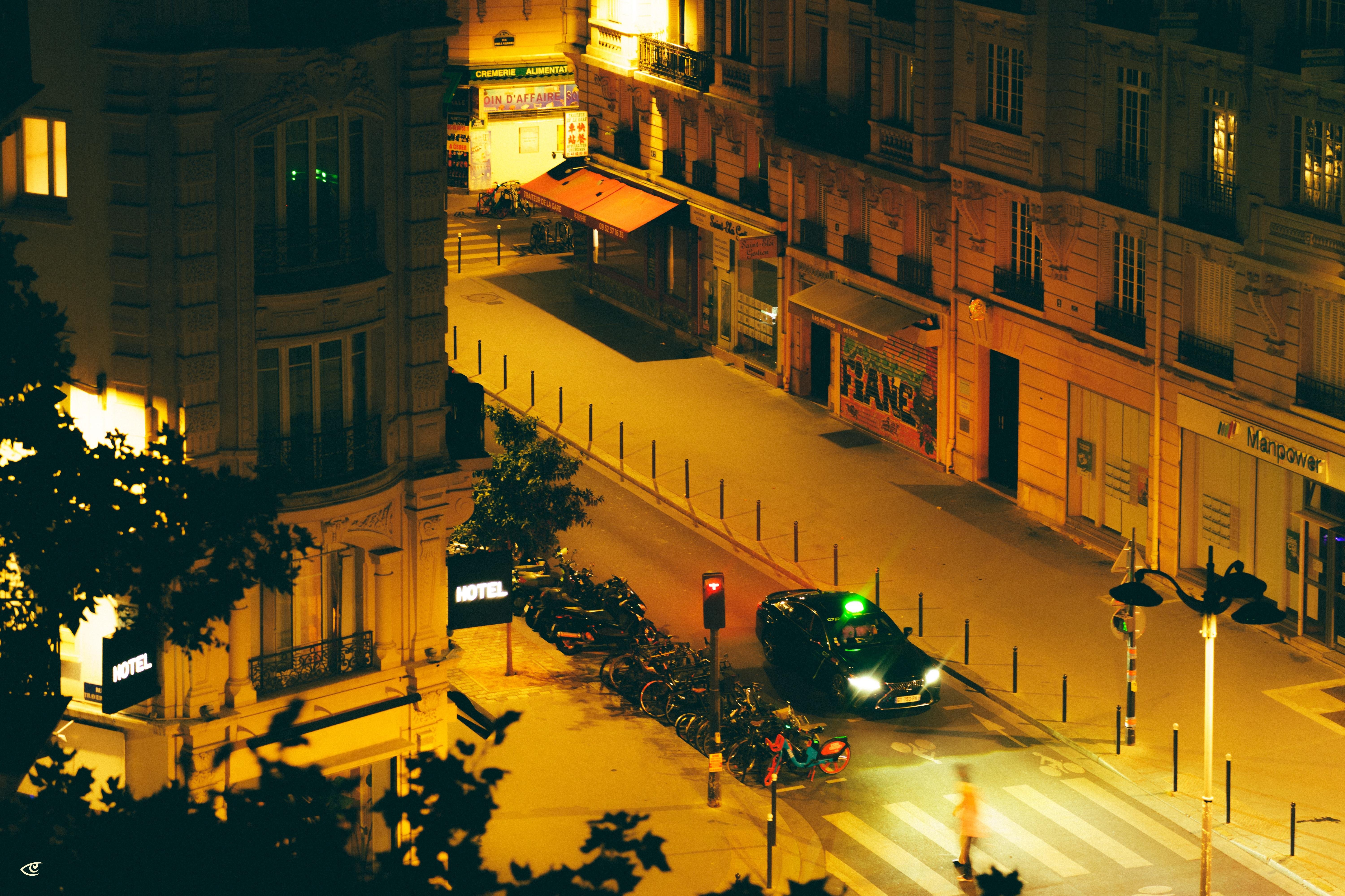 Elevated nighttime view of a city intersection with a zebra crosswalk, a taxi stopped at the curb, rows of parked motorcycles, and warm-lit buildings, including a corner hotel sign and storefronts along an empty street.