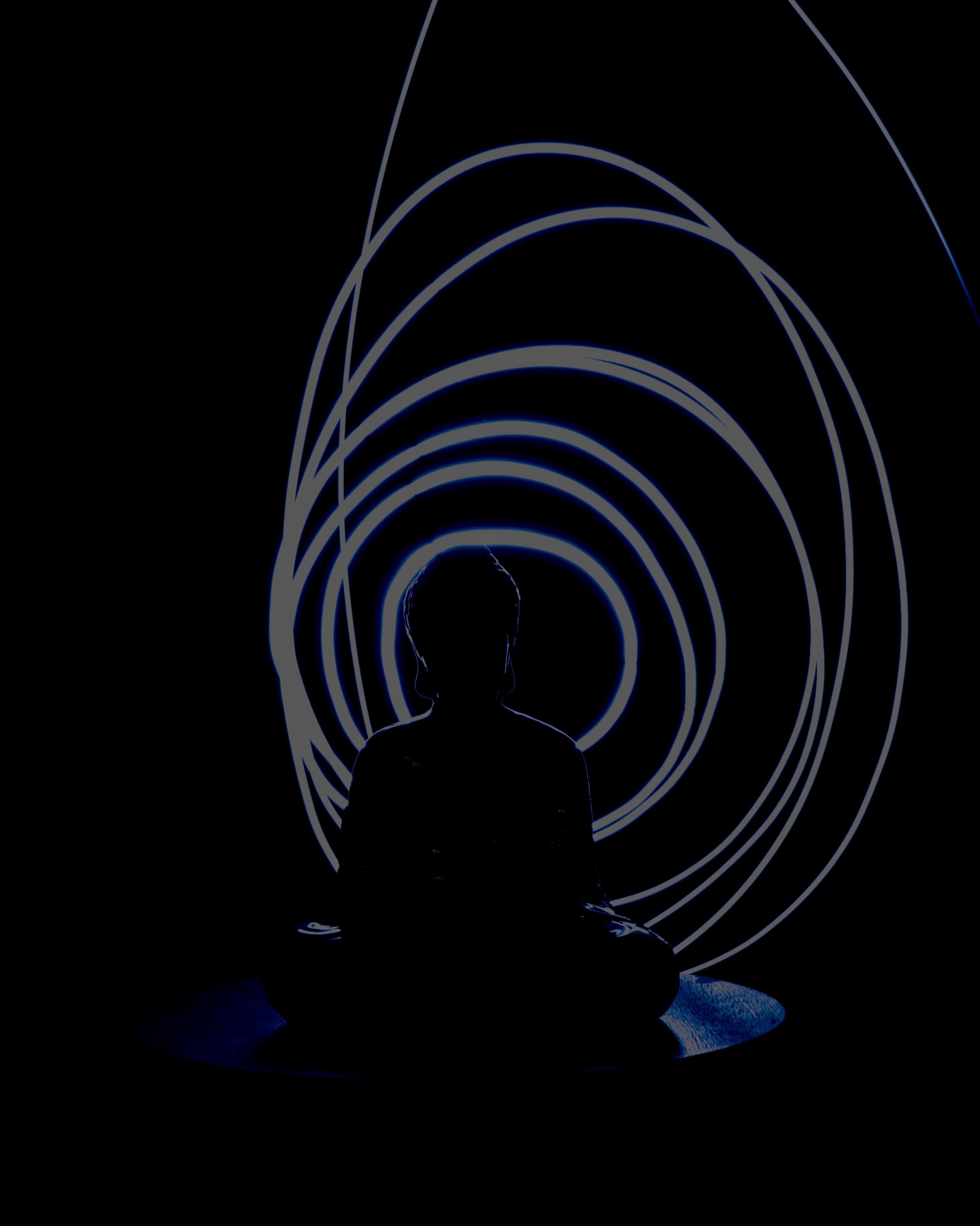The image shows a silhouette of a person sitting cross-legged, likely in a meditative pose. It is a buddha. Behind him, there are concentric rings of light, creating a visually striking backdrop. The overall scene suggests themes of meditation, peace, and tranquility.