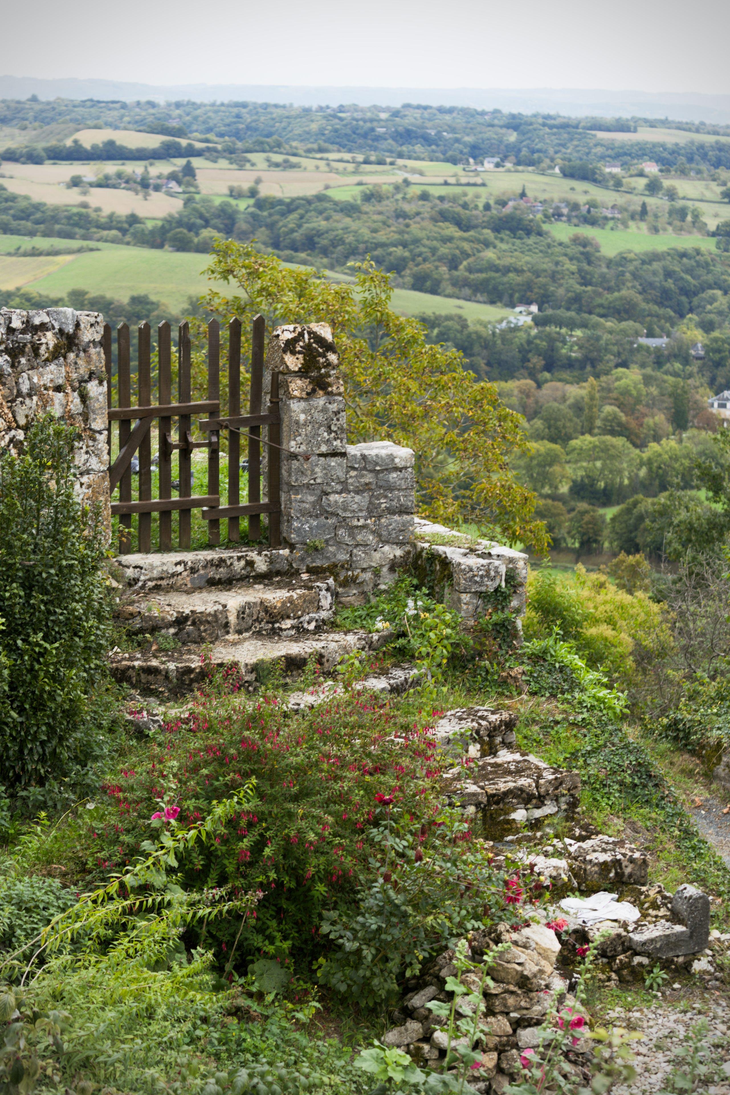 Photo format paysage montrant des escaliers en très vieilles pierre, couvertes de mousse et de lichen, menant à un portail en métal protégeant un mur en ruine. En arrière plan, un paysage menant champs et forêts