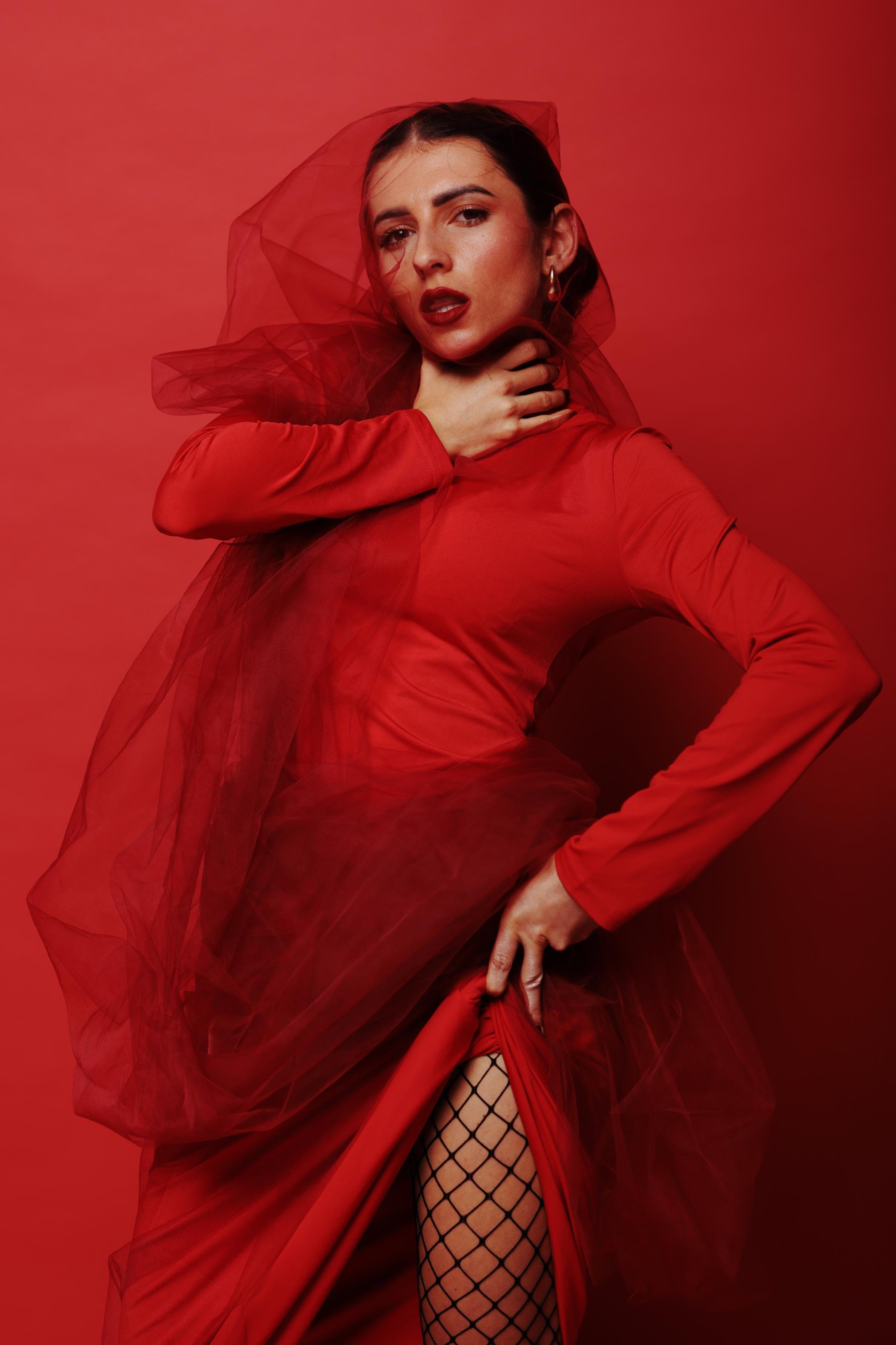 Woman wearing a red dress and veil against a matching red background. She holds her neck with one hand and lifts her dress with the other, revealing fishnet stockings. Her makeup includes bold red lipstick and small hoop earrings.