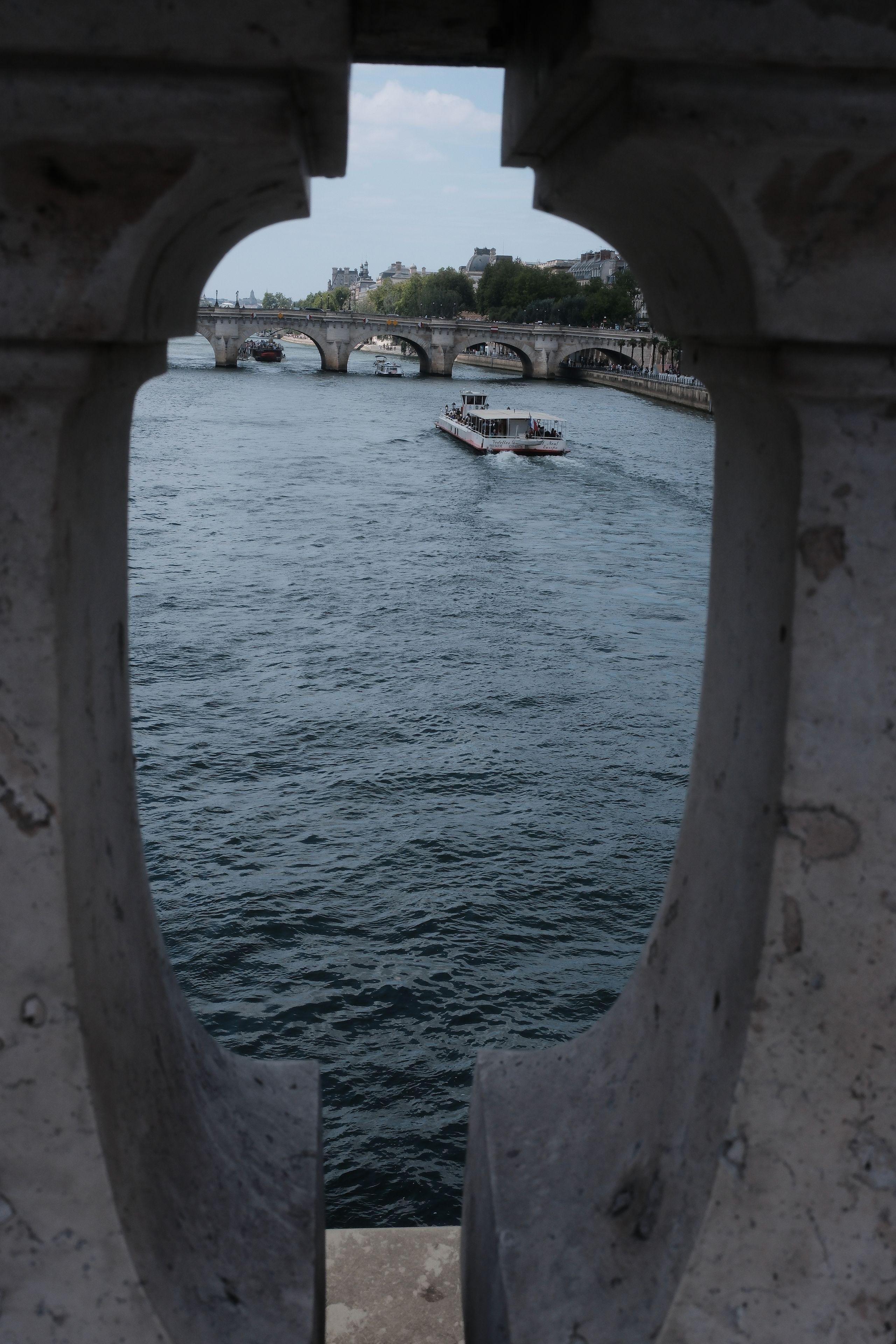 Photo format portrait, montrant un bout de la Seine avec un bateau allant au loin, le tout cadré par la rambarde en pierre d’un pont, entre 2 barreaux en pierre