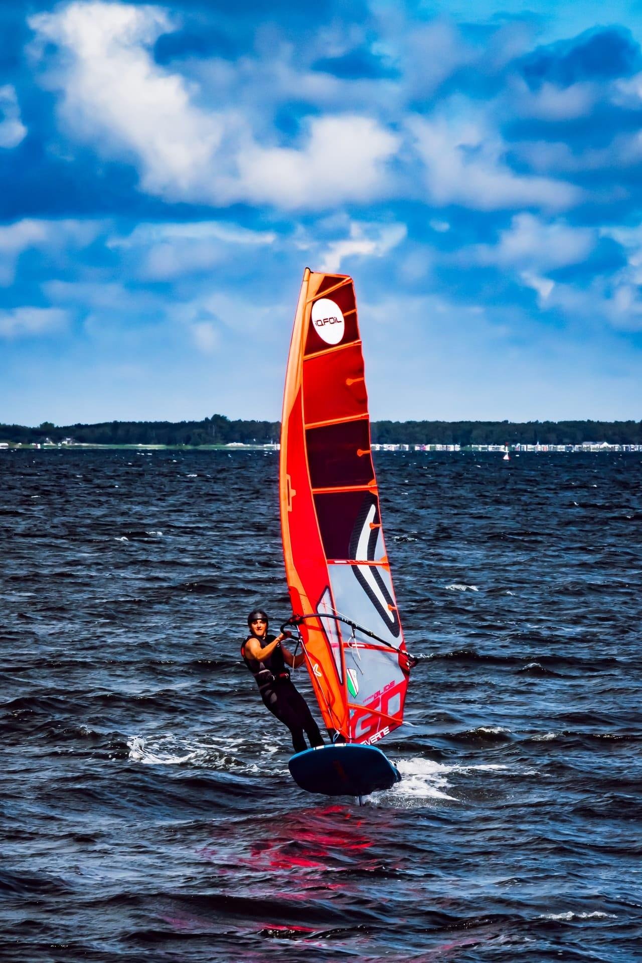 Windsurfer in a black wetsuit riding a board on choppy water, holding a tall red-and-orange sail with a white circular logo; distant shoreline along the horizon under a blue sky with large clouds.