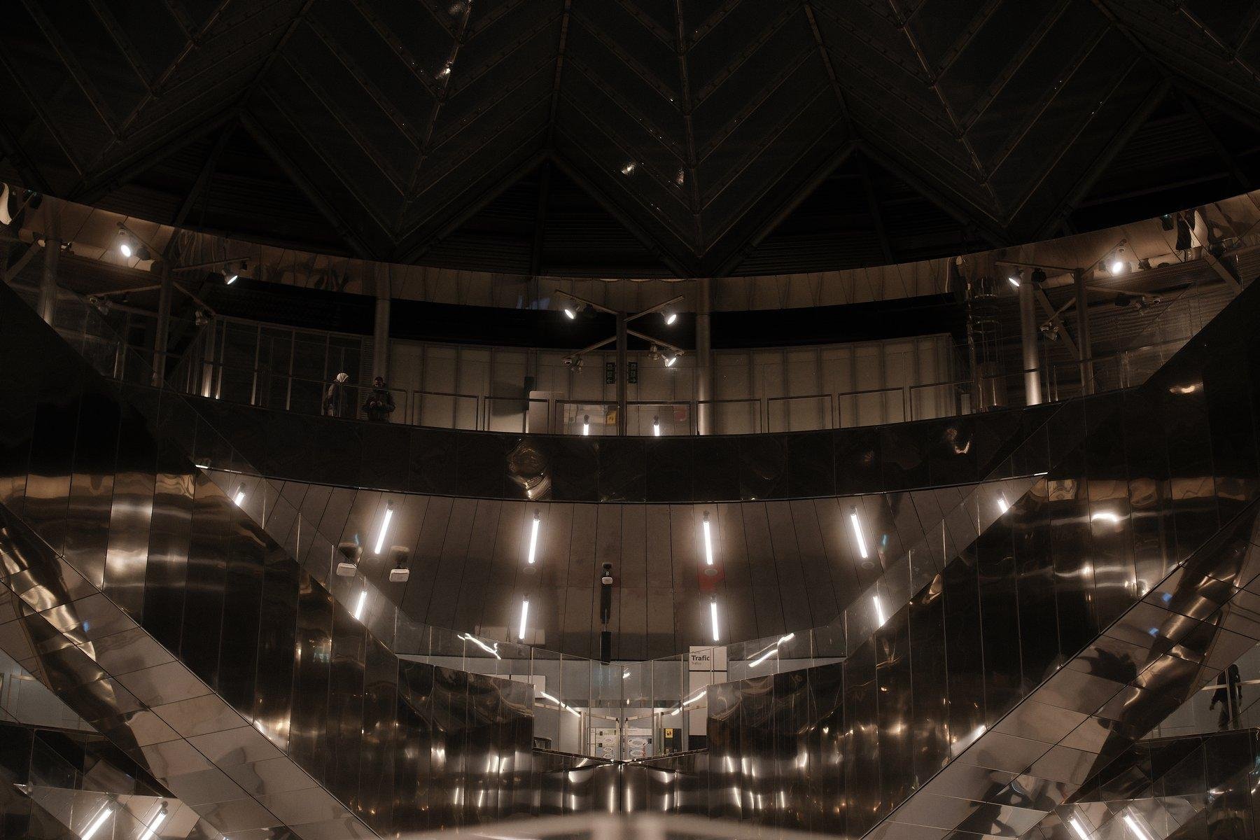 Photo format paysage couleur, montrant l’intérieur de la station de métro Villejuif Gustave Roussy, qui est tout de métal réfléchissant et une coupole en verre. Deux escalators de profil forment un V symétrique.
