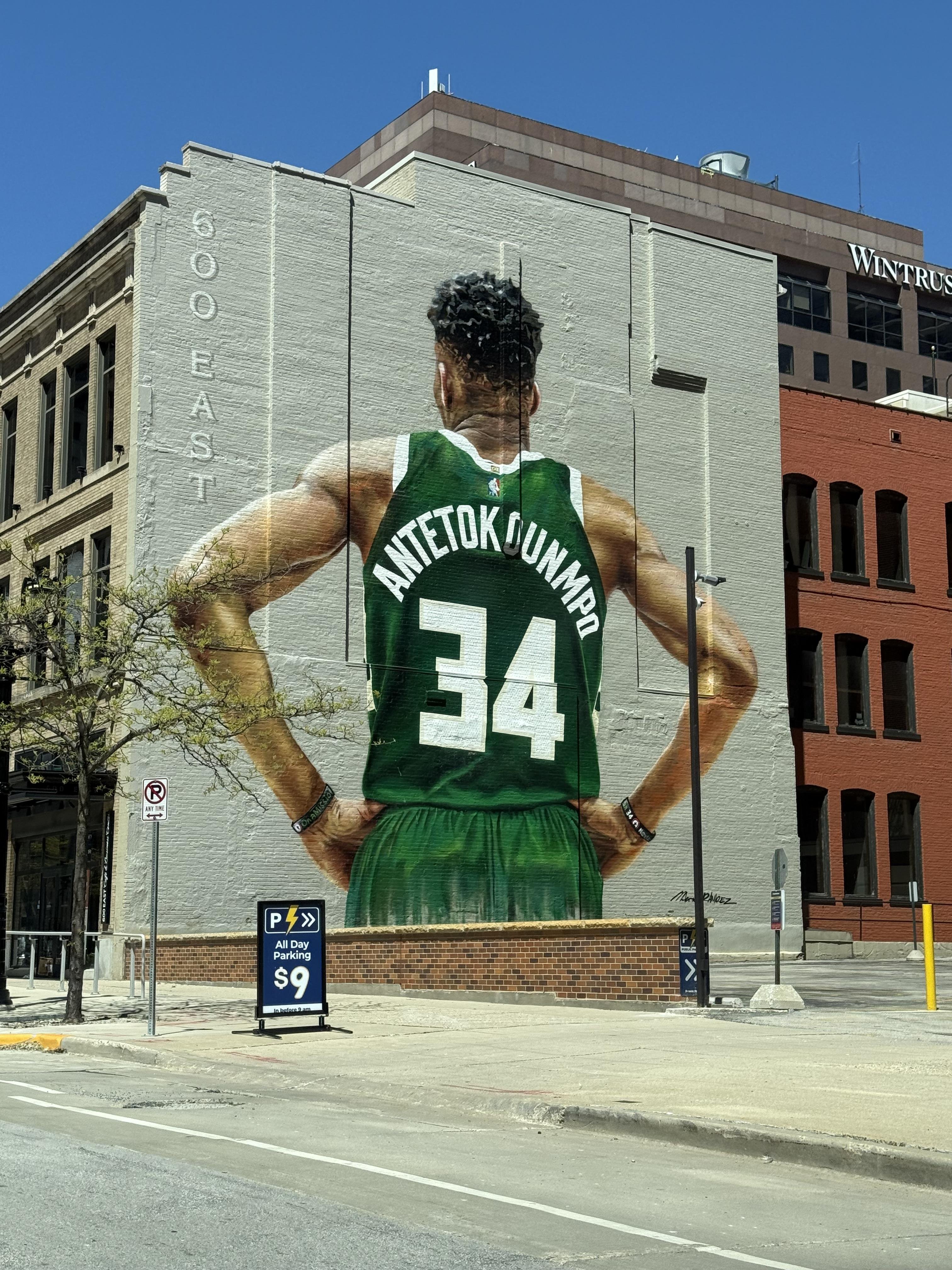Large mural on a building's side depicting a basketball player wearing a green jersey with the name "Antetokounmpo" and number 34, facing away with hands on hips. Adjacent streets and a parking sign advertising $9 all-day parking are visible. Nearby buildings include a reddish-brown structure with "Wintrust" signage. The location is marked with "600 East" on the building. Clear blue sky above.