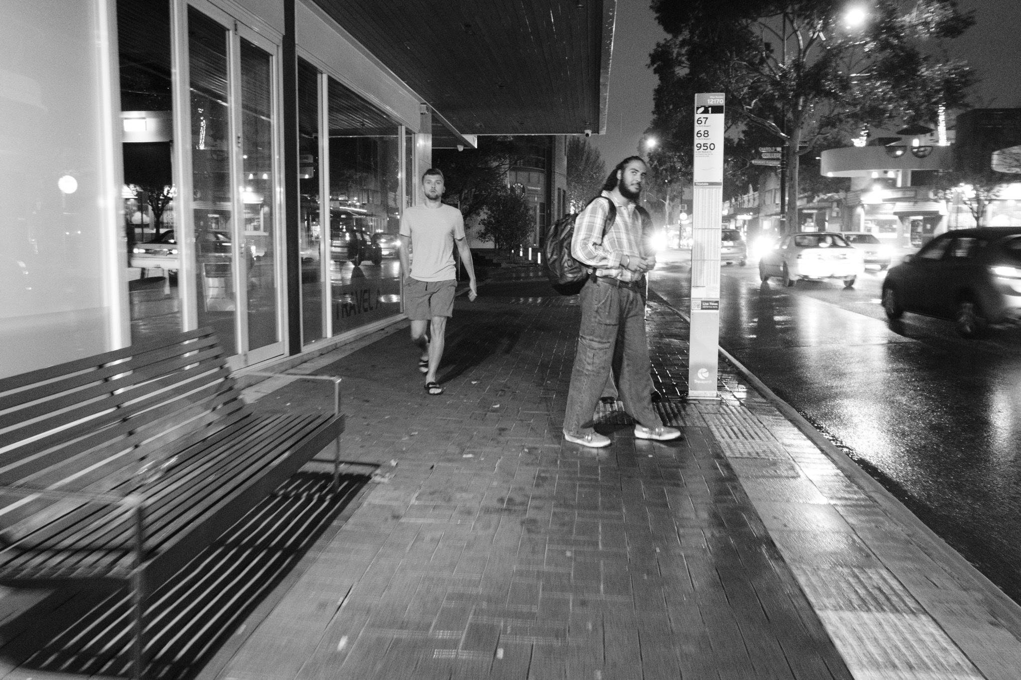 Black-and-white nighttime street scene with a wet sidewalk beside a road with passing cars; two pedestrians near a bus stop sign, a bench and glass storefront on the left, and streetlights reflecting on the pavement.