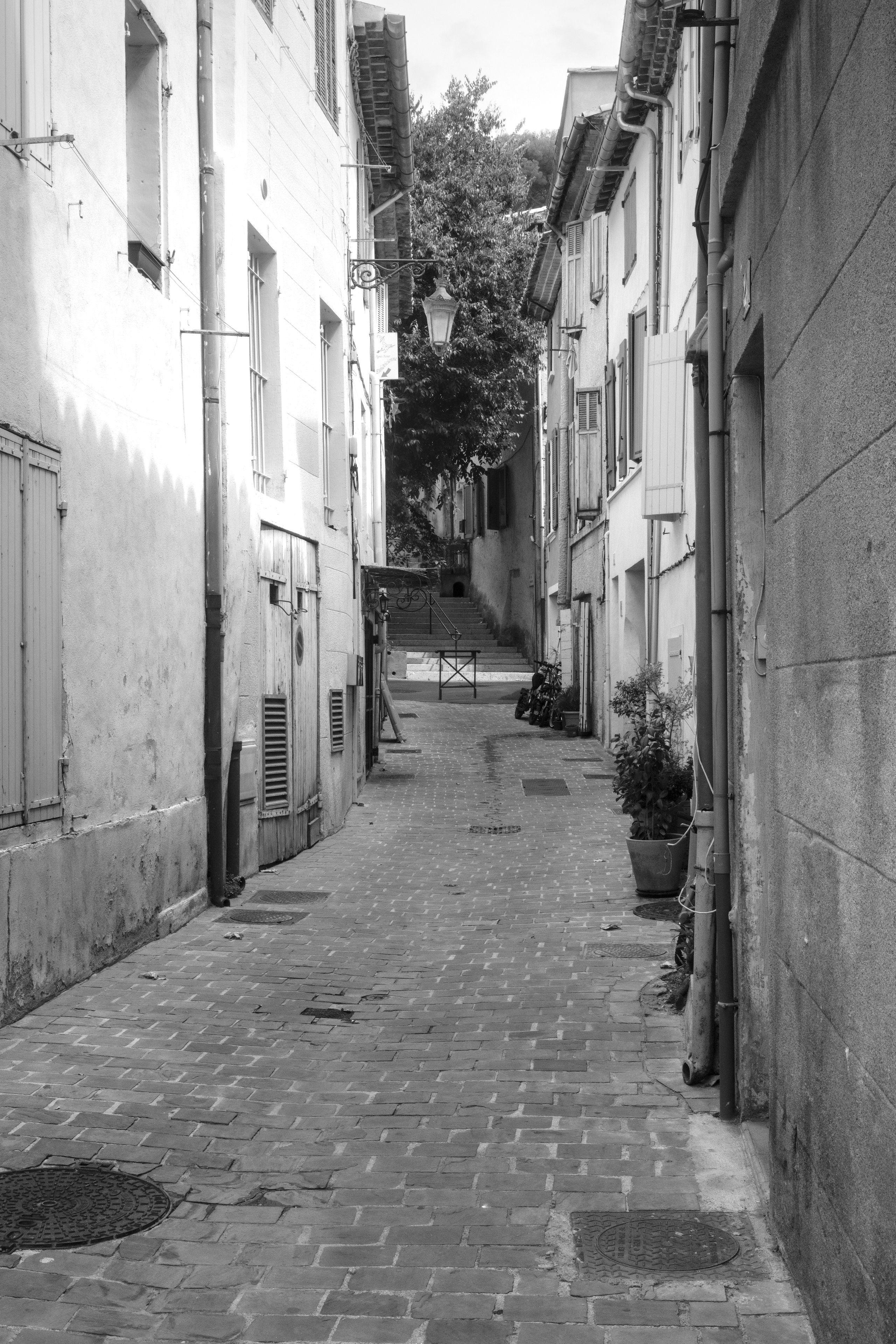 Photo portrait, en noir et blanc, montrant une ruelle piétonne pavée entourée de petites maisons de ville, qui monte vers des escaliers au loin