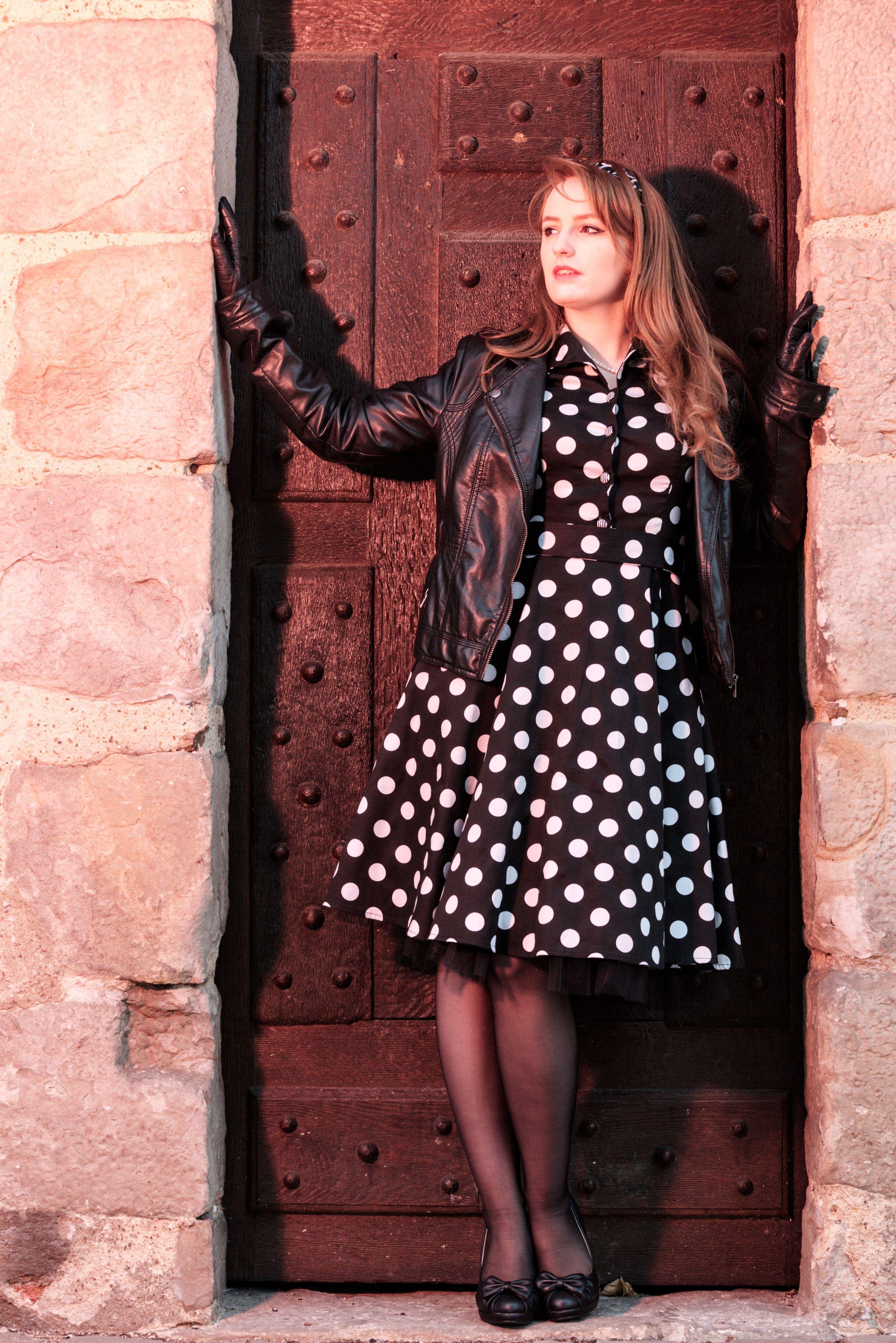 Photo format portrait, montrant une jeune femme rousse au cheveux long dans l’encadrement d’une vieille porte en bois. Elle est coiffée d’un bandeau noir et blanc attaché dans le style années 50. Elle porte une robe noire à gros pois blanc qui s’arrête aux genoux, un blouson en cuir, des gants noir en cuir et des chaussures noires avec un nœud sur leur dessus. Elle a les mains posée de sur chaque côté du cadre de porte, jambes serrées, penchée sur sa gauche en repliant le bras, tête tournée vers sa droite et regardant au loin. La scène est éclairée par une lampe orange/rose