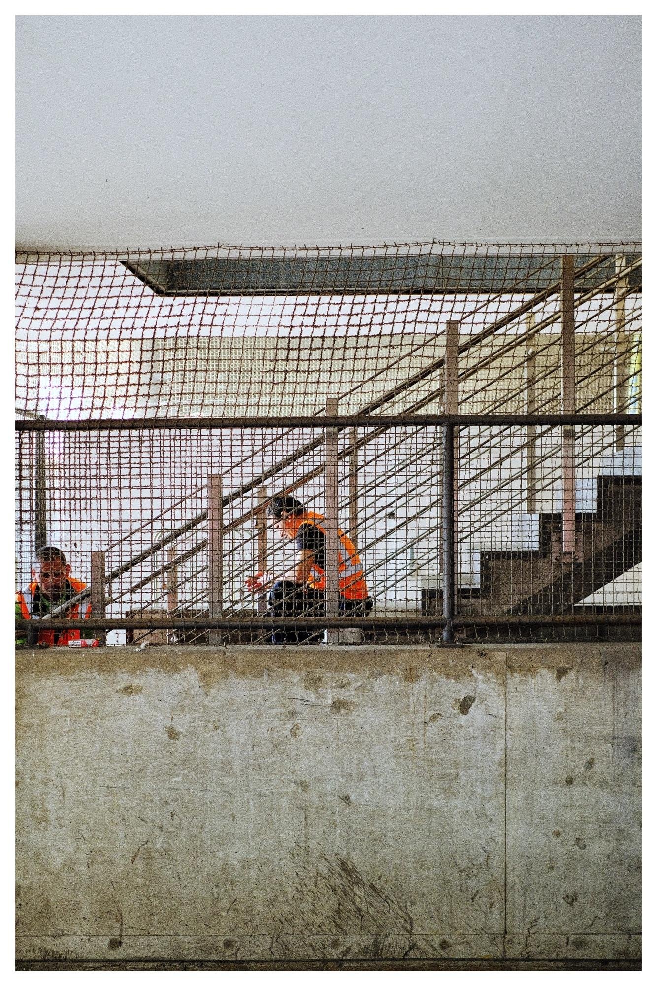 Two workers in orange safety vests behind a wire-mesh fence on an elevated concrete platform, with a metal staircase and handrails in the background.