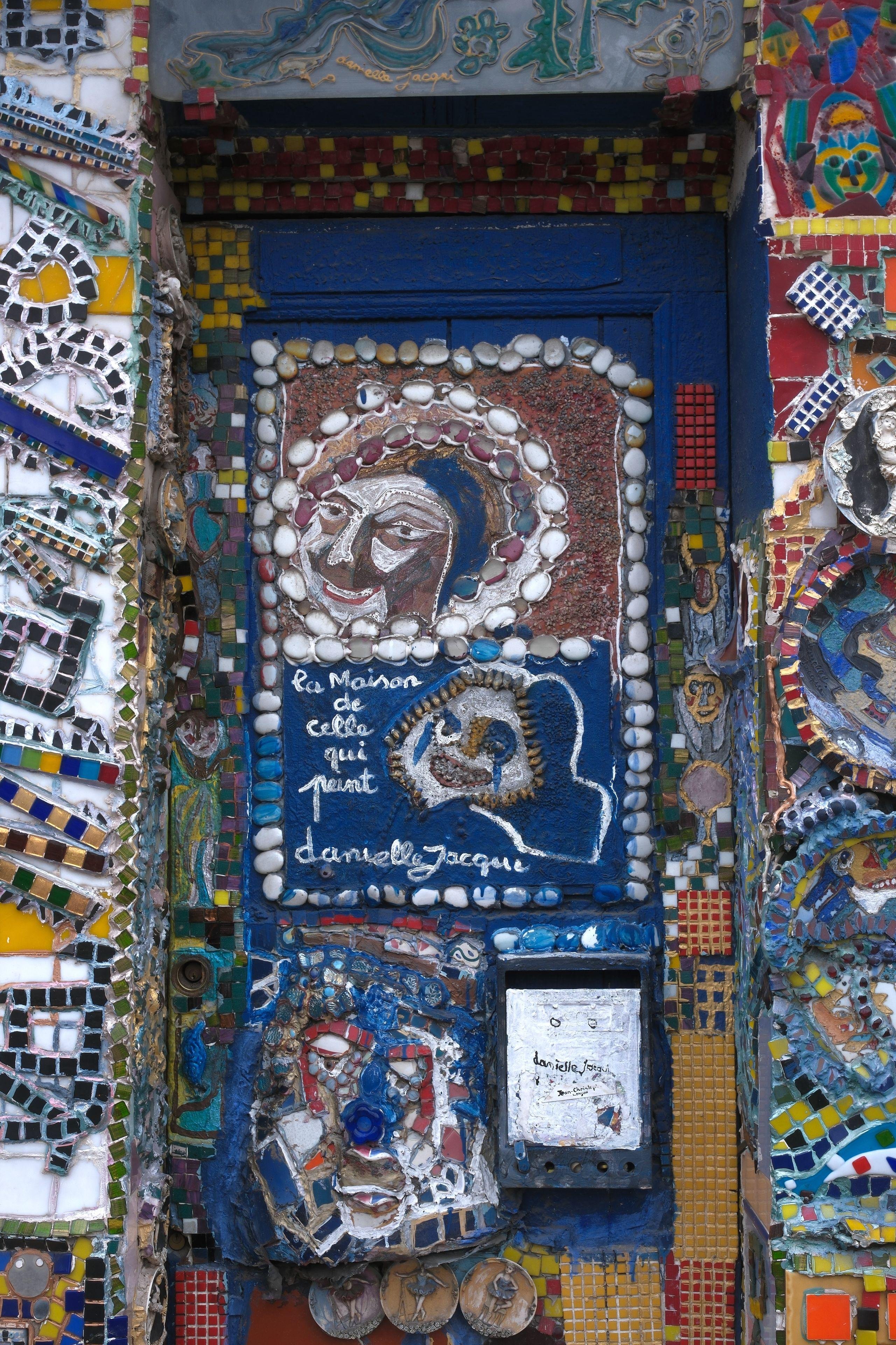 A door really heavily decorated with pebbles, paint, ceramic, random object, all stuck in a more or less random fashion, with sometime patterns of human face or animal ones