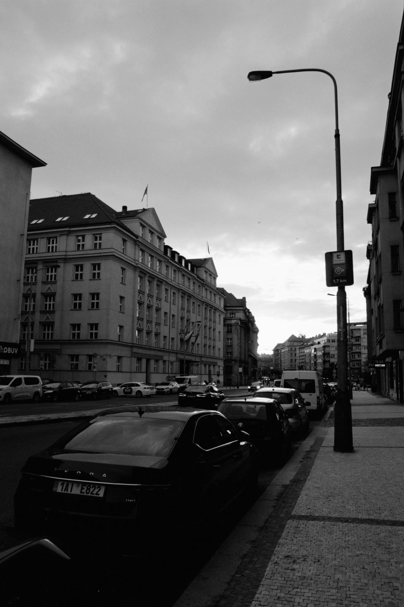 Black-and-white street scene with parked cars along a curb beside a tiled sidewalk, multi-story buildings lining both sides of the road, and a tall streetlight with a transit sign in the foreground under a cloudy sky.