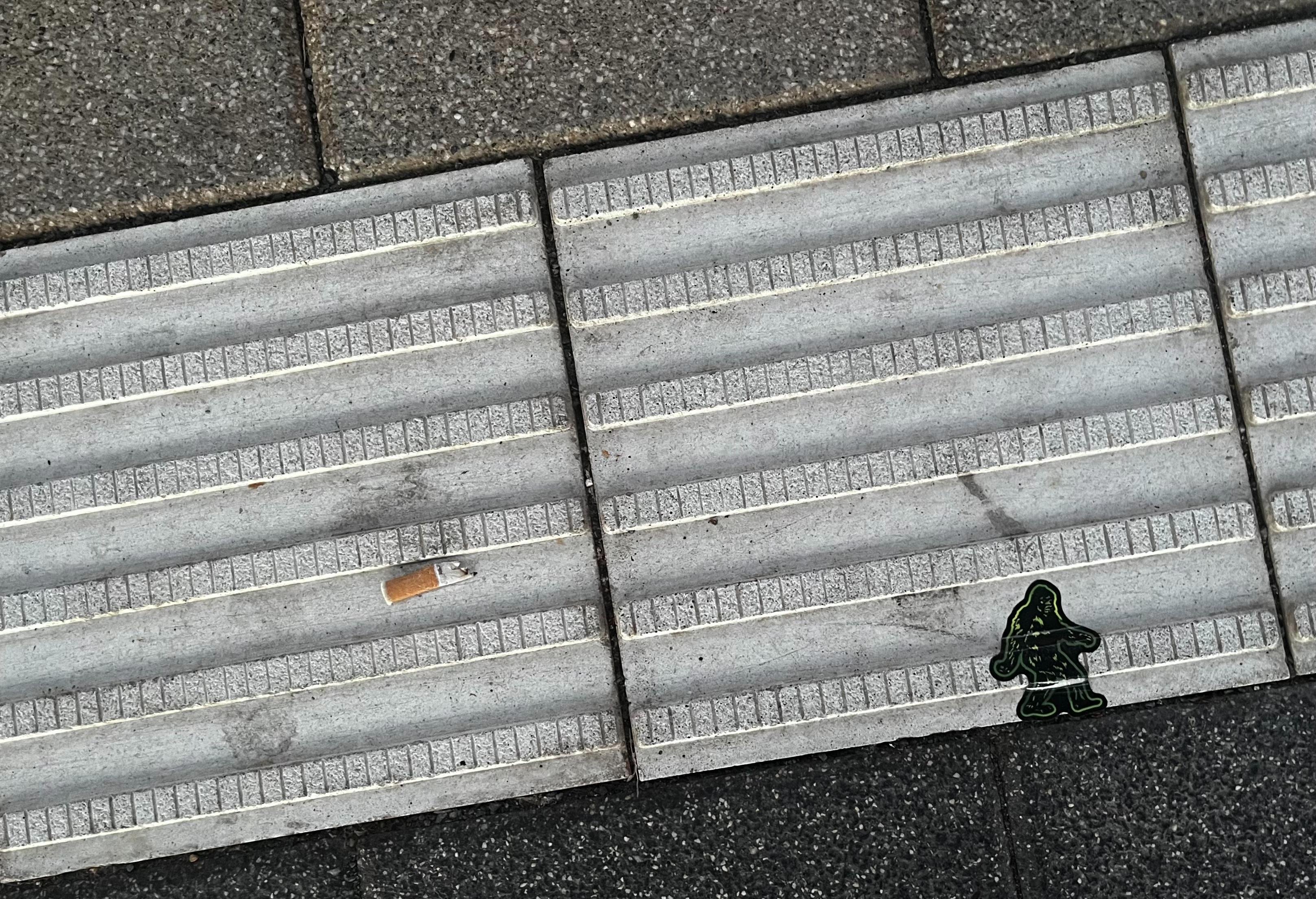 Closeup view of a train platform floor gfrom straight above with two ribbed stone plates bordered by dark paving stones, a cigarette butt on the left plate, and a small green figure sticker near the lower right, positioned at the edge, as if it was ascending a hillside / slope.