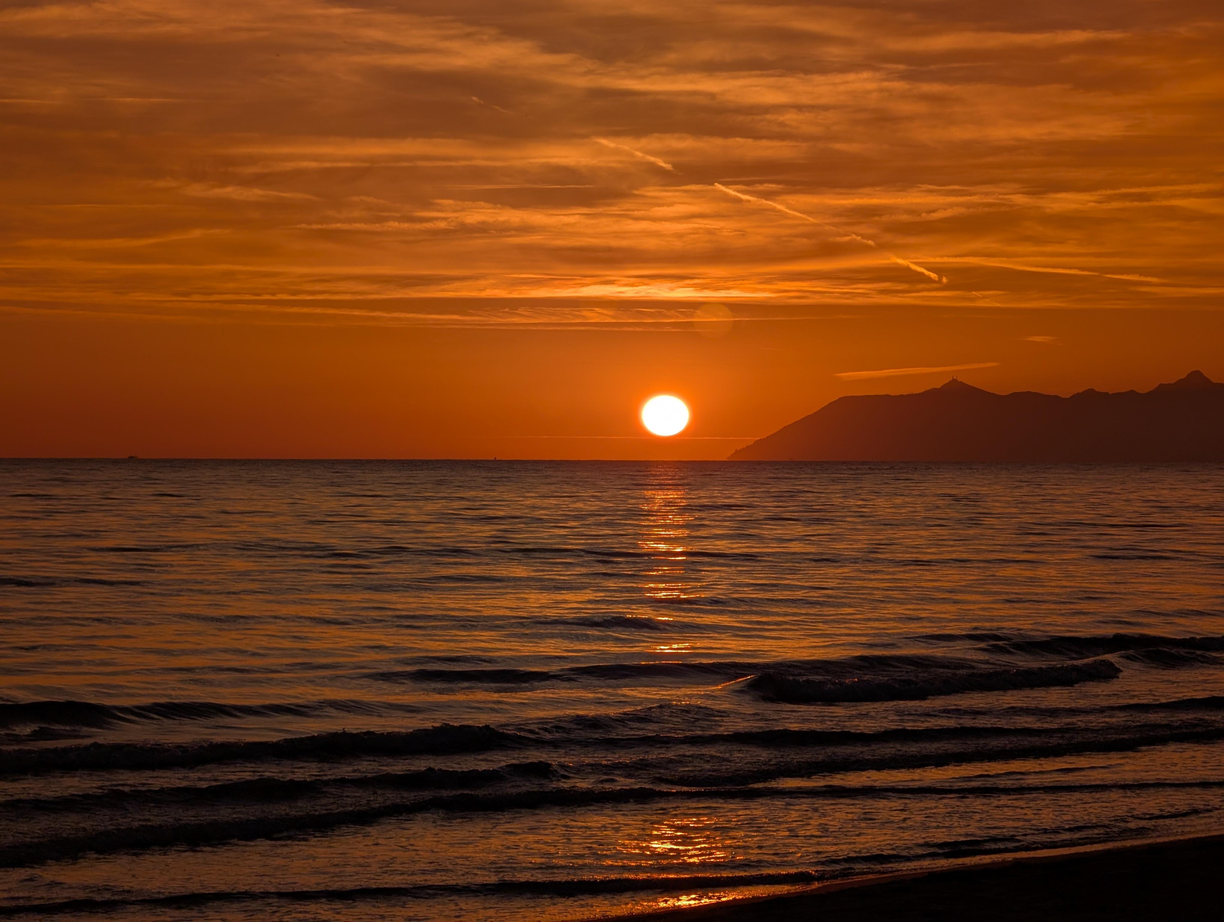 The image shows a beautiful sunset over the ocean. The sun is low on the horizon with an orange sky and some clouds. There are gentle waves on the water, and a silhouette of a distant landmass or mountain can be seen on the right side. It creates a serene and picturesque scene.