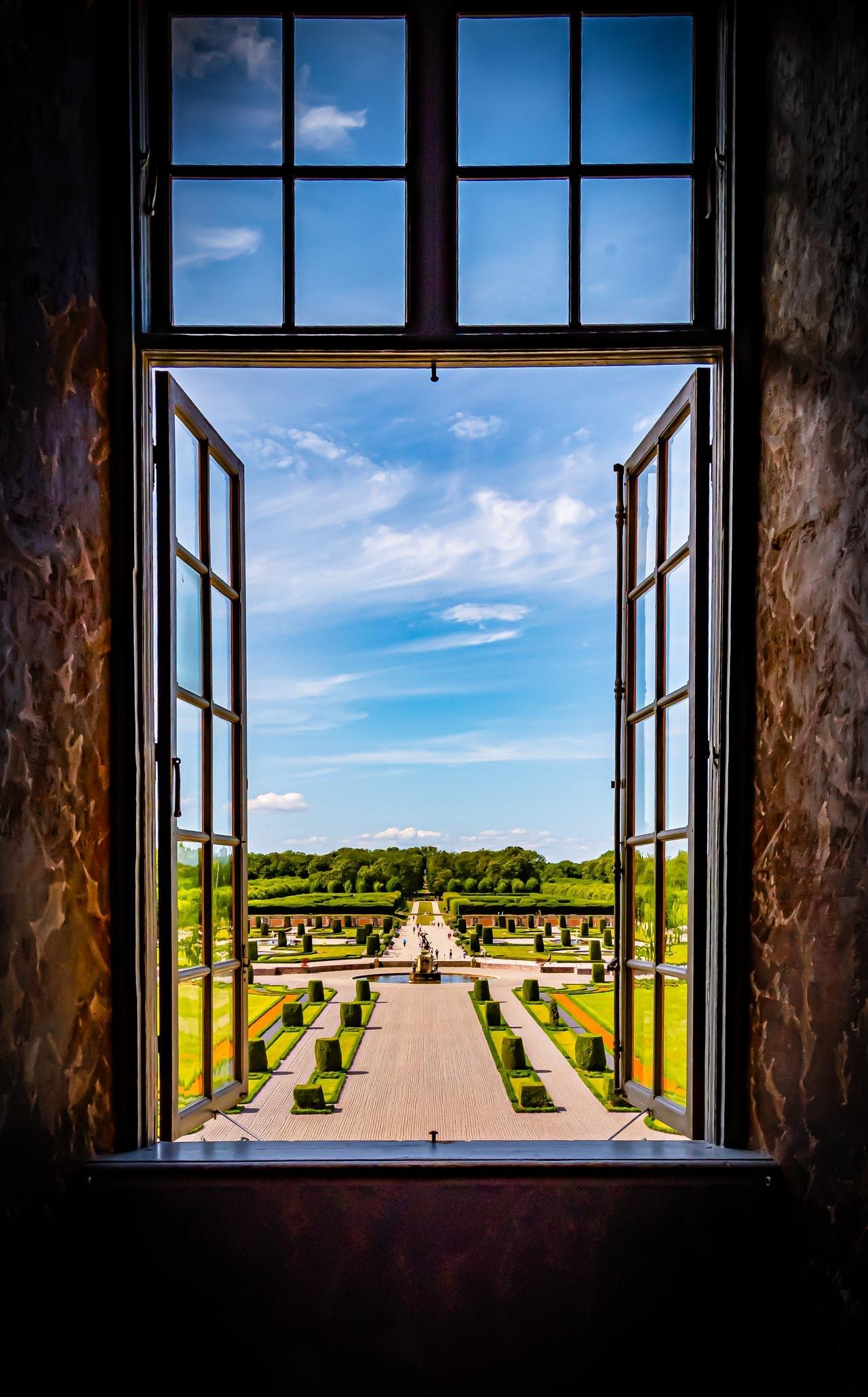 Open window frame overlooking a formal garden with symmetrical pathways and neatly trimmed hedges. A wide path leads to a distant fountain. The sky is bright blue with scattered white clouds. Trees line the garden's edge in the background.