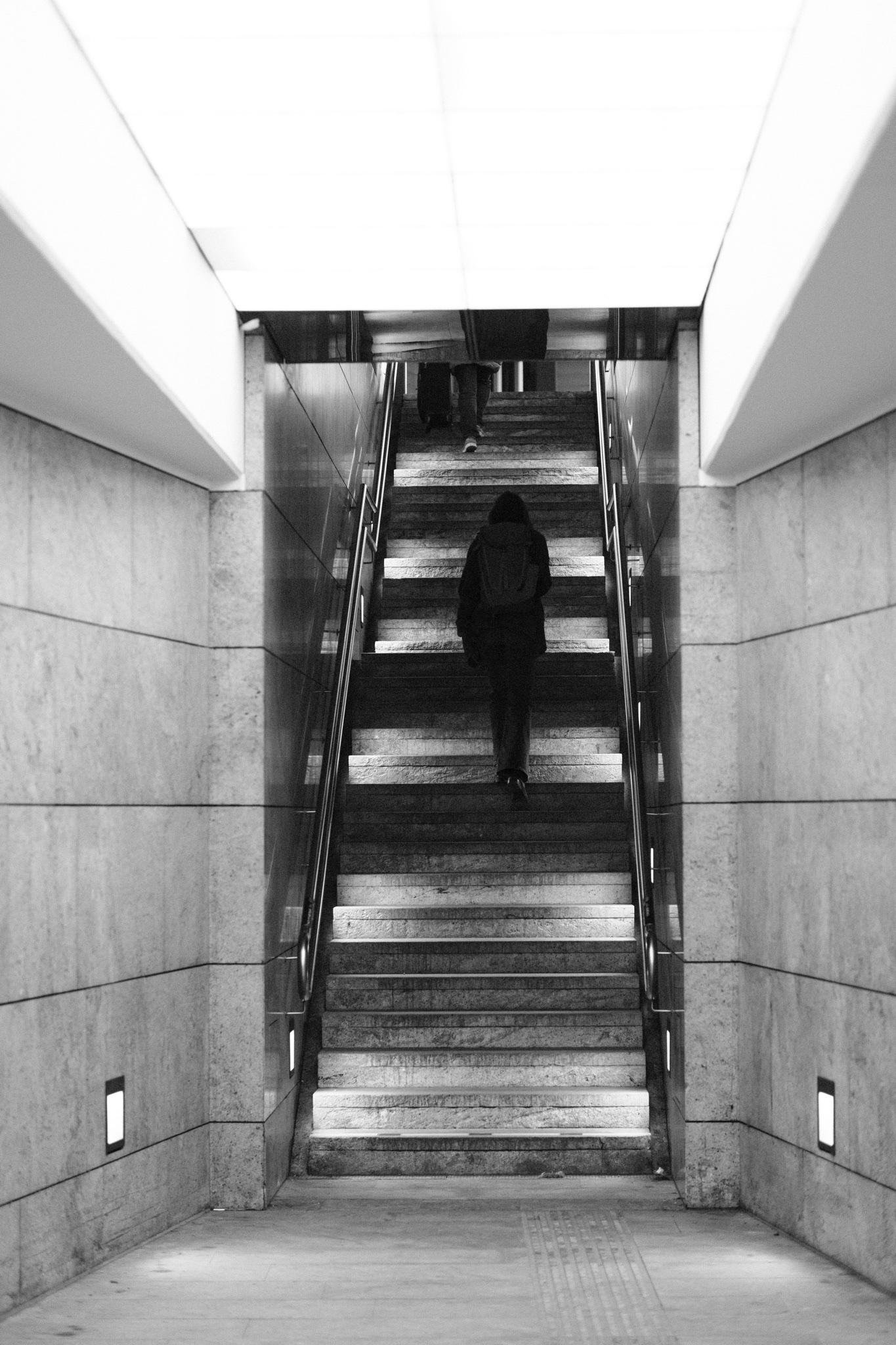 Black-and-white view up a wide stairwell in an underground passage, with metal handrails and tiled walls; a person with a backpack climbs the stairs while another figure is near the top under bright ceiling lights.