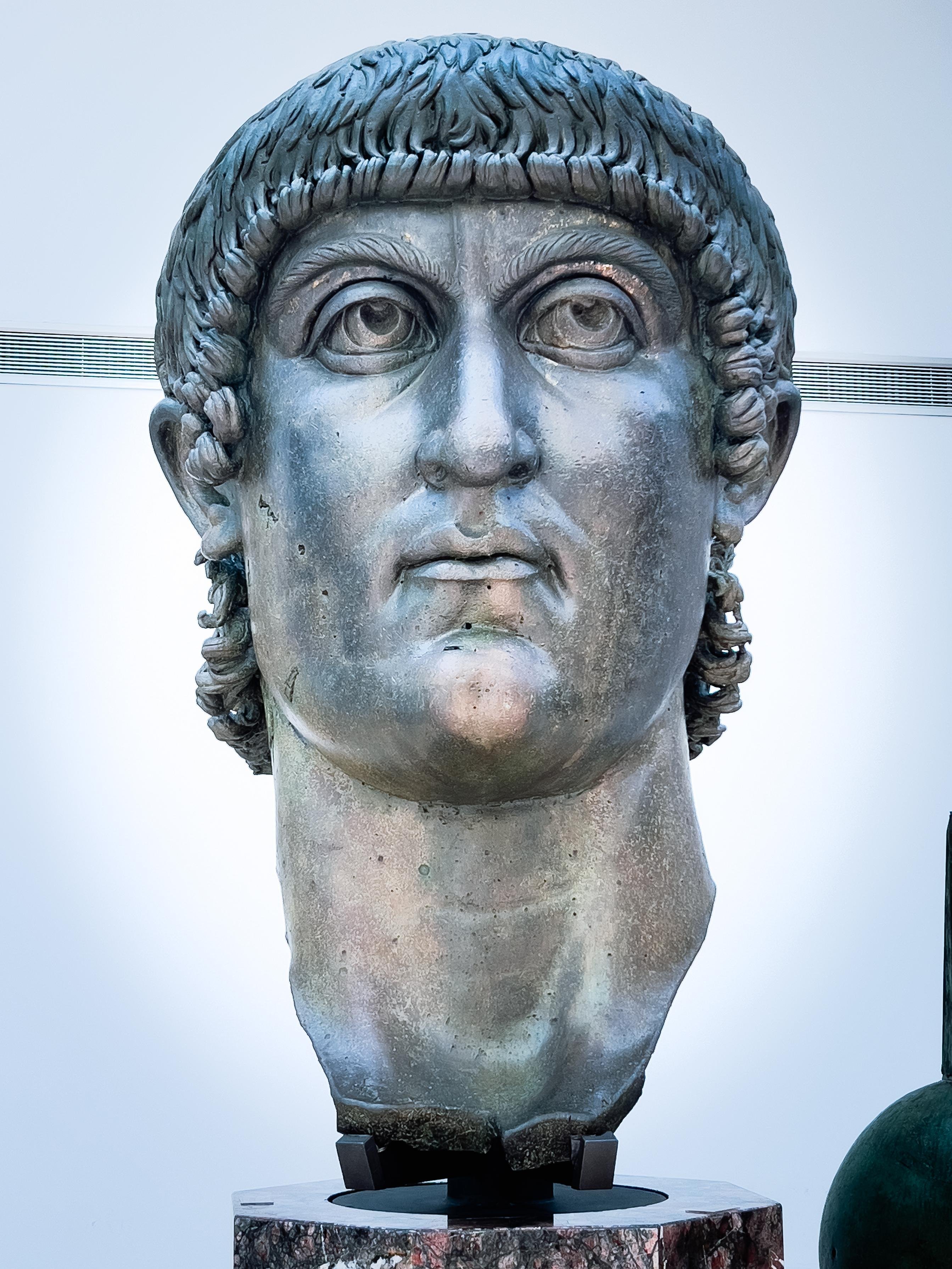 Grande testa in marmo di una statua, probabilmente raffigurante un imperatore romano. Lo stile e i dettagli suggeriscono che sia un importante artefatto storico, possibilmente da una statua famosa come il Colosso di Costantino. È montata su una base per l'esposizione.