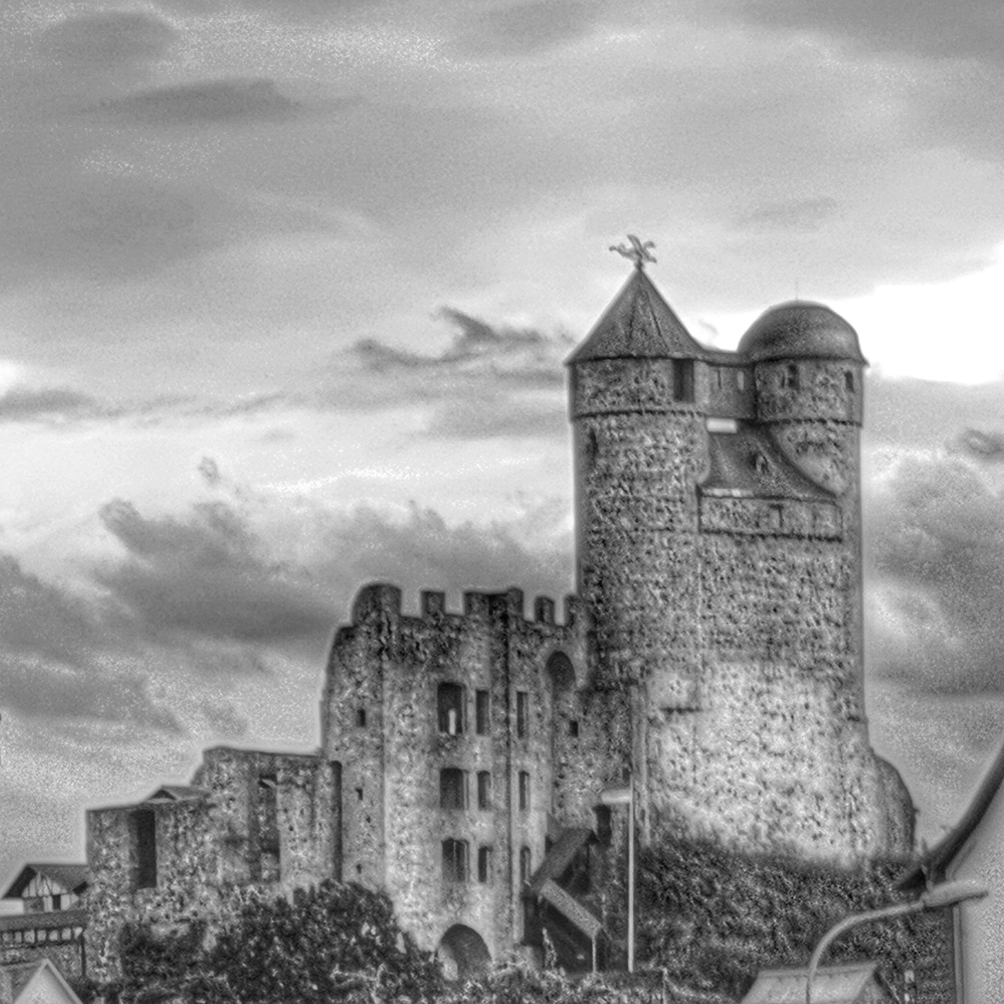 A black-and-white, slightly grainy photograph shows Burg Greifenstein, a medieval hilltop castle in Hesse, Germany. The imposing stone structure dominates the scene, rising against a dramatic clouded sky. At the right stands the castle’s iconic double-tower silhouette: a tall cylindrical tower with a pointed roof topped by a decorative cross, connected to a second domed tower by a short, fortified walkway. To the left, the massive ruins of the main keep display arched openings, weathered walls, and uneven battlements that hint at centuries of history. Dense foliage surrounds the base of the castle, with a few rooftops from nearby houses visible in the foreground. Burg Greifenstein dates back to the 12th century and is known for its twin-towered profile and the Glockenwelt (Bell Museum) housed within today. The monochrome effect emphasizes texture, age, and the castle’s enduring medieval character.