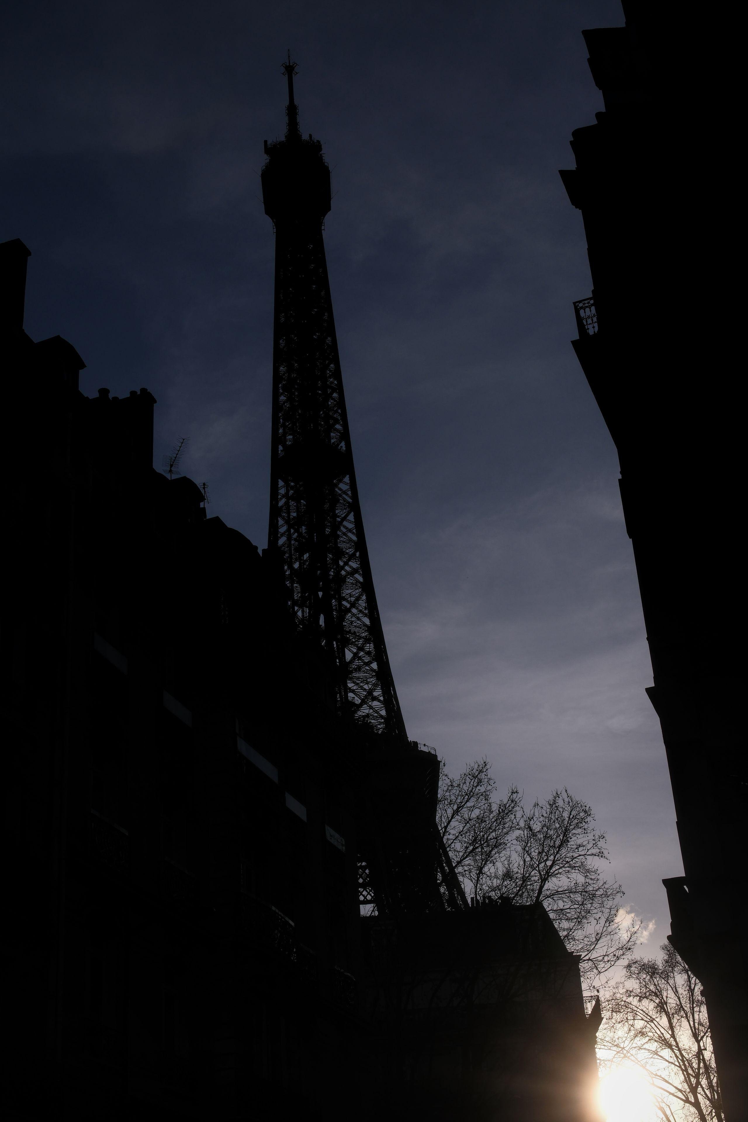 Photo verticale. Entre 2 rangée d’immeubles de par et d’aurte de l’image, la silhouette du haut de la tour Eiffel dépasse sous un ciel sombre, en contre-jour à cause du soleil en bas à droite de l’image