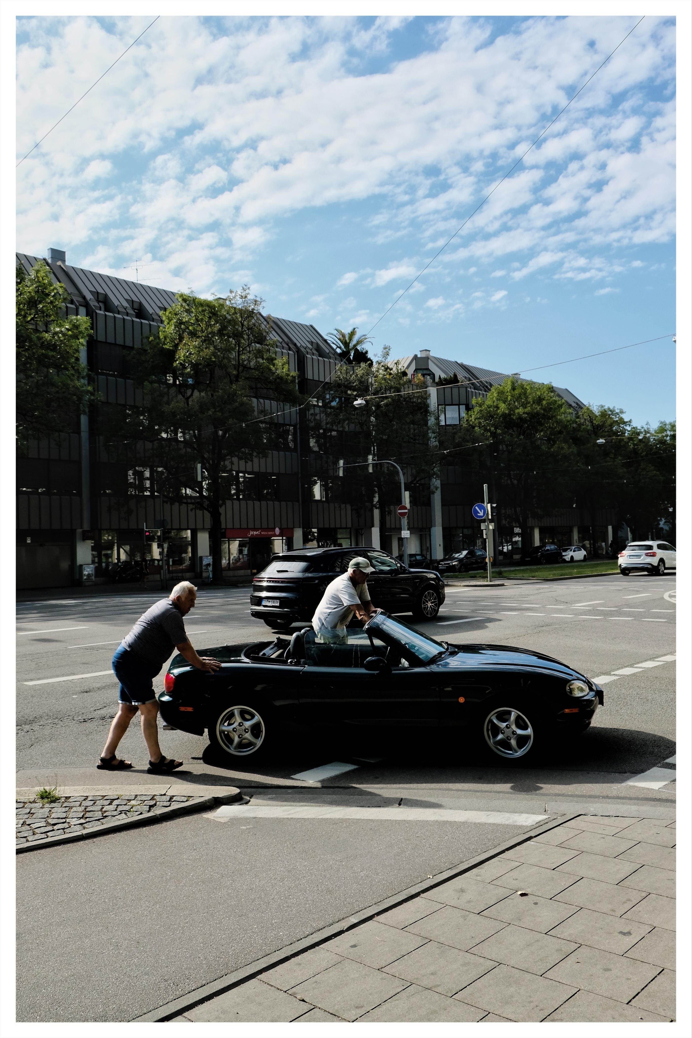 Two men push a black convertible stopped at a street intersection, with modern dark-roofed buildings, trees, traffic signs, and other cars under a partly cloudy sky.