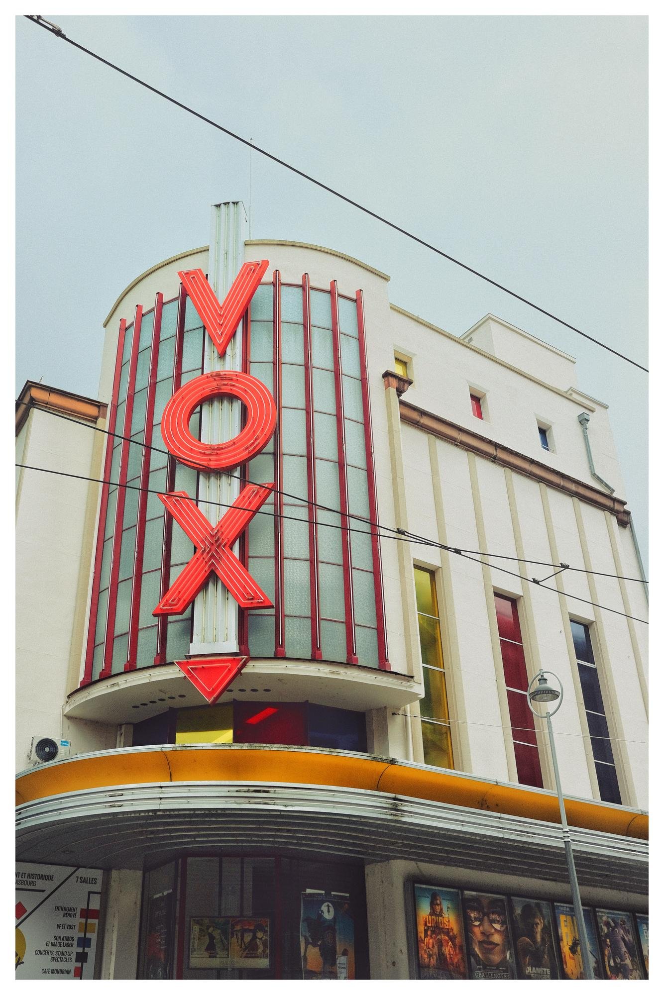 Art deco cinema facade with a large vertical red neon “VOX” sign on a rounded glass-front tower, overhead power lines crossing the sky, and movie posters displayed along the entrance below.