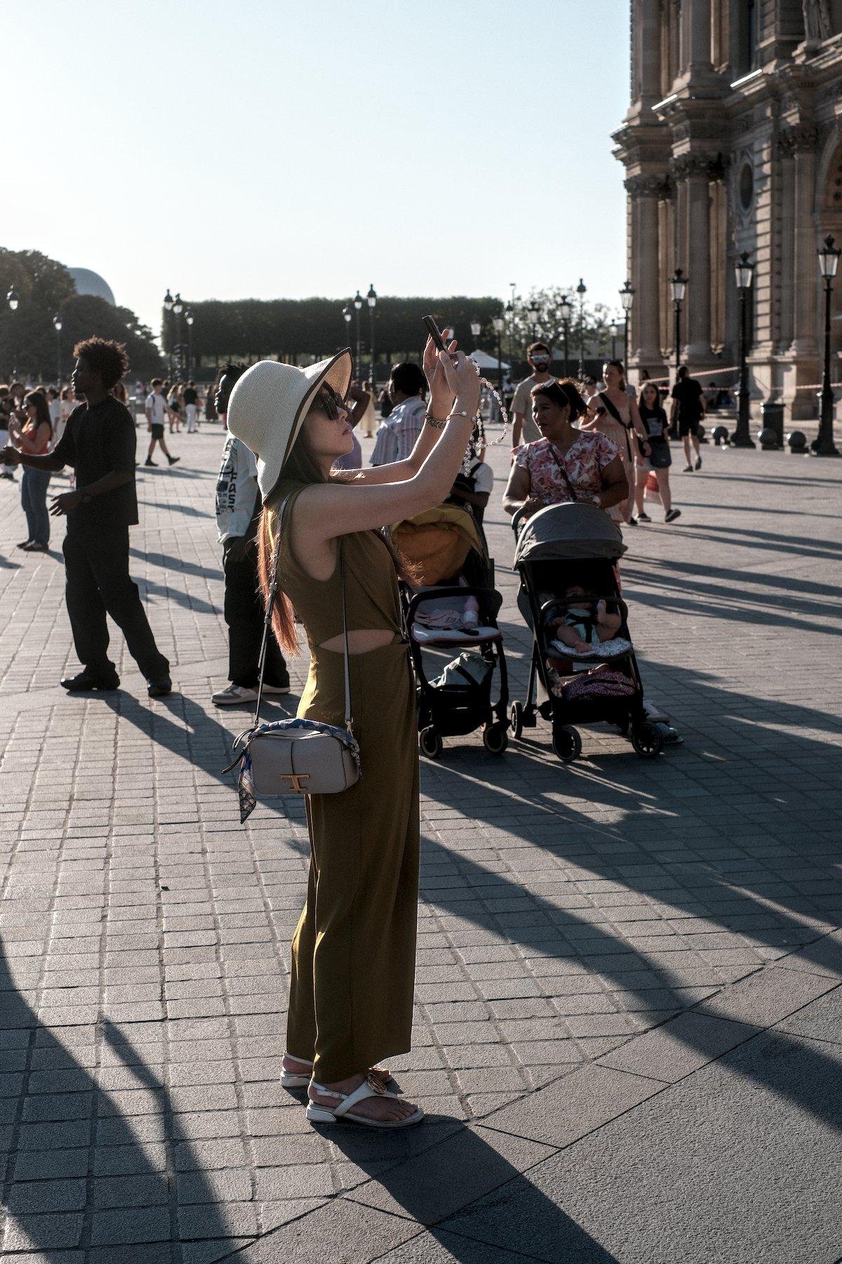 Photo verticale. Sur le parvis du Louvre, une femme en haut marron sans manche, pantalon léger assorti, chaussures ouvertes blanches, lunettes de soleil, chapeau beige clair, est en train de prendre une photo avec son téléphone