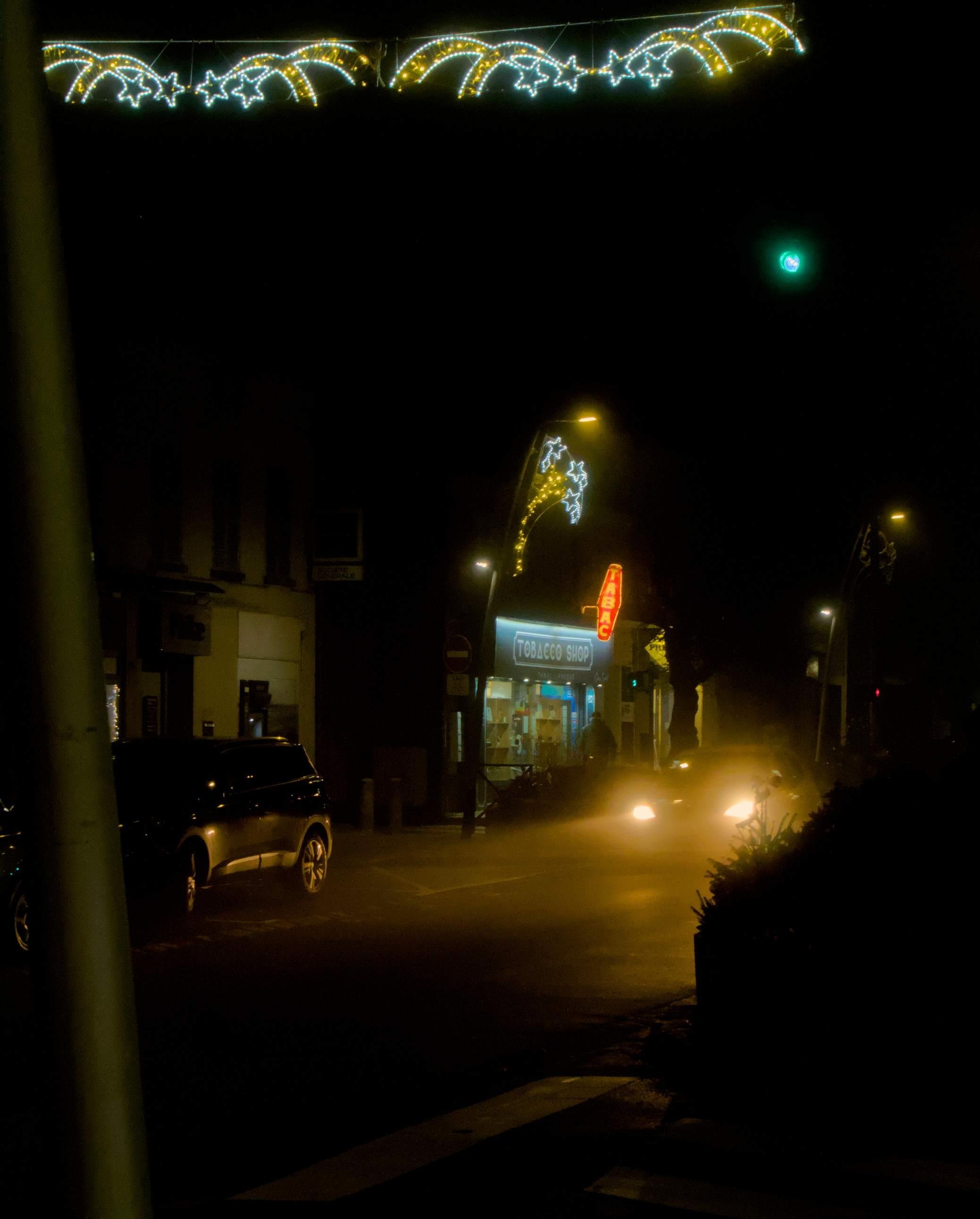 La photographie dépeint une scène urbaine de nuit avec des décorations lumineuses de noël en forme d'étoiles suspendues en l'air et accrochées à un lampadaire. En dessous, il y a une boutique avec une carotte de buraliste et une enseigne bleue disant "Tabacco shop". Devant la devanture, des phares de voiture de couleur ambre éblouisse l'objectif. Au premier plan, à gauche, un poteau longe la hauteur de l'image. Un passage piéton, un marquage de stop et des jardinières de rues décorent le coin inférieur droit de l'image.
