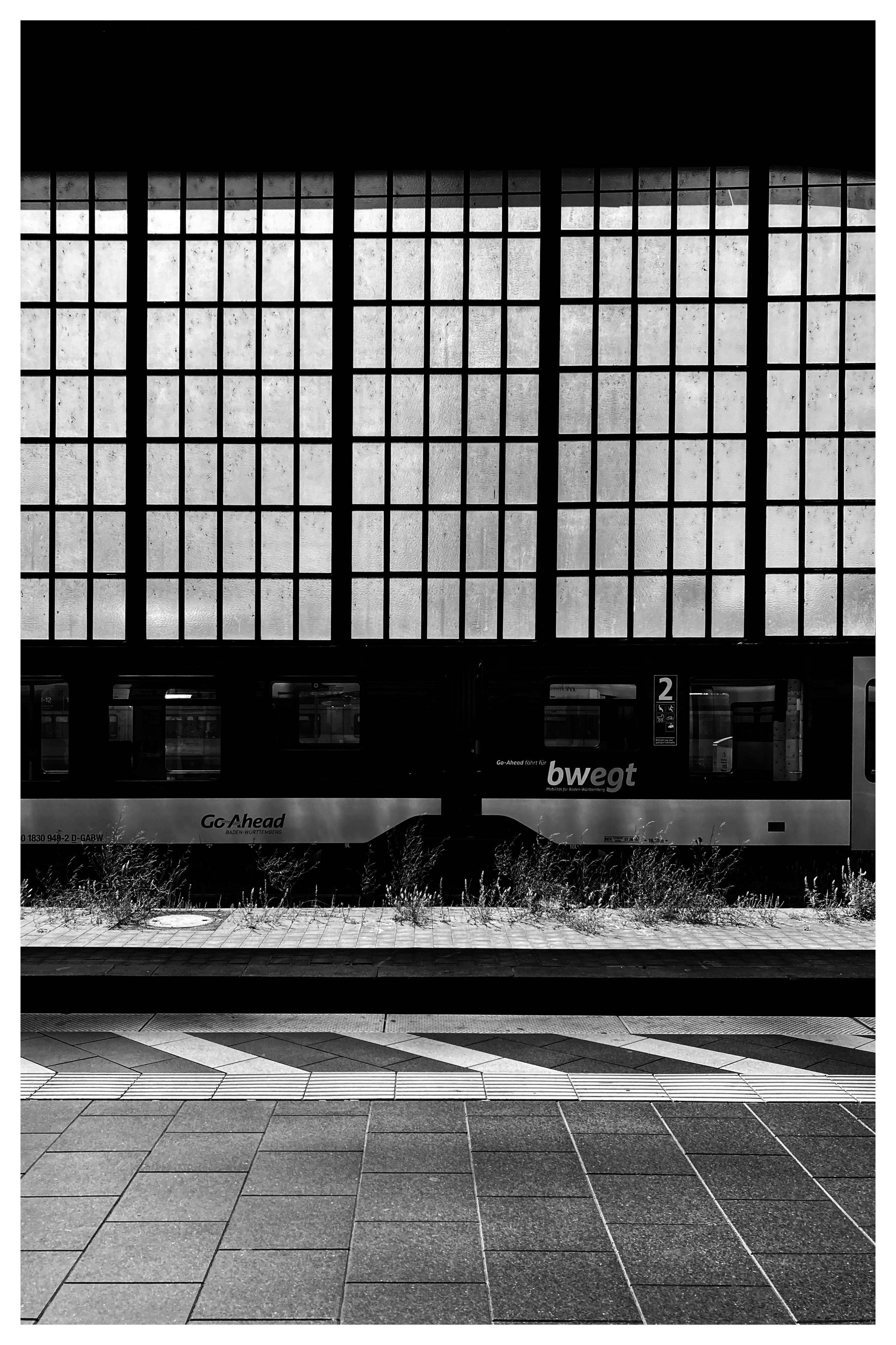 Black-and-white view of a train at a station platform in front of large grid-patterned windows; tiled pavement with diagonal markings in the foreground, weeds along the track edge.