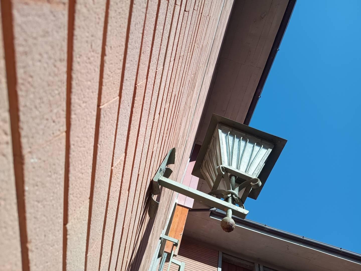 Inquadratura dal basso di un lampioncino su un muro di mattoni, la parte destra della foto è ampiamente occupata dal cielo azzurrisimo.