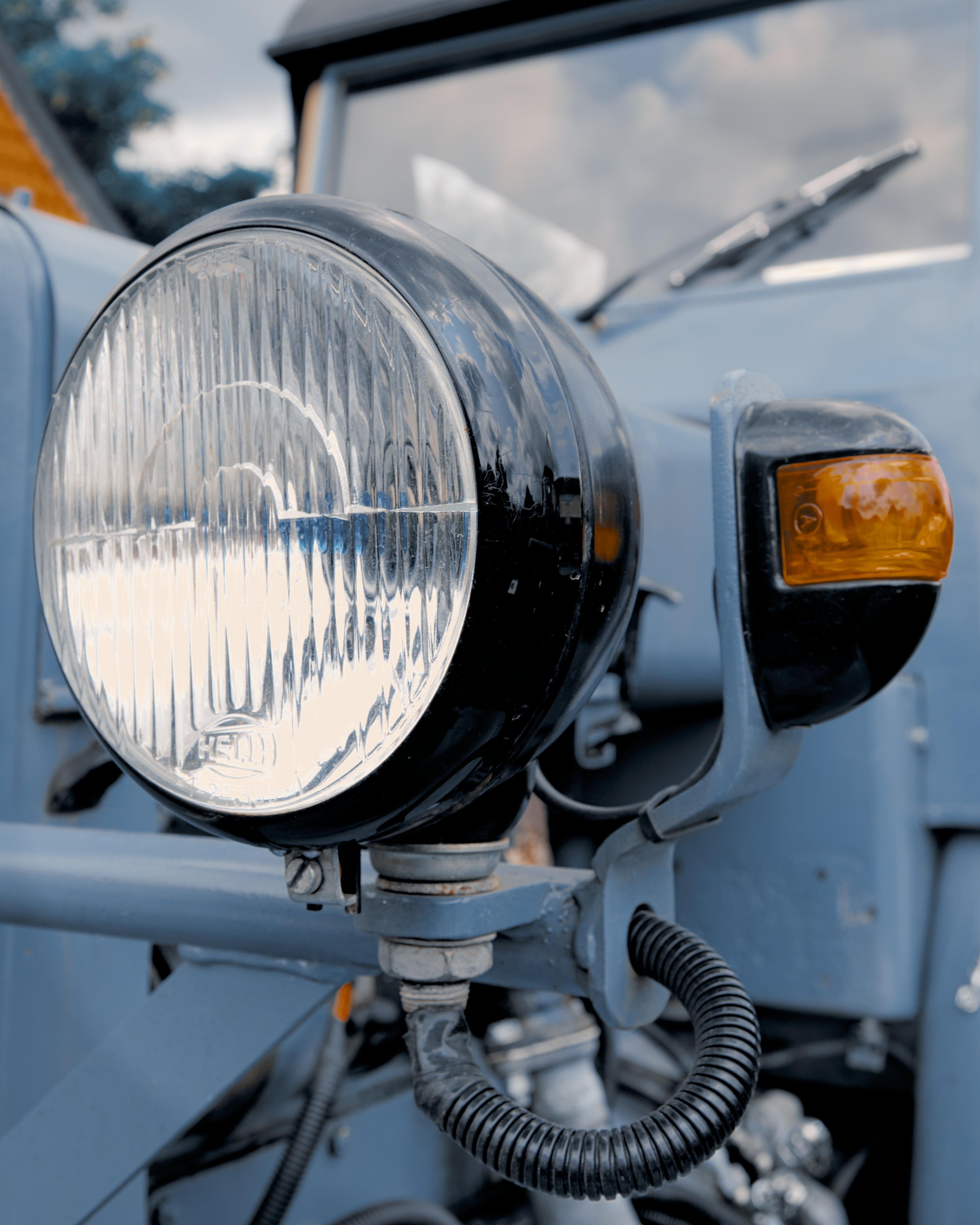 Close-up of the front headlight and blinker of a vintage blue vehicle. The large, round headlight features a textured glass surface, while the smaller amber blinker is mounted above it on the right. Power cables and mounting brackets are visible, with parts of the vehicle’s frame and windshield wipers in the background.