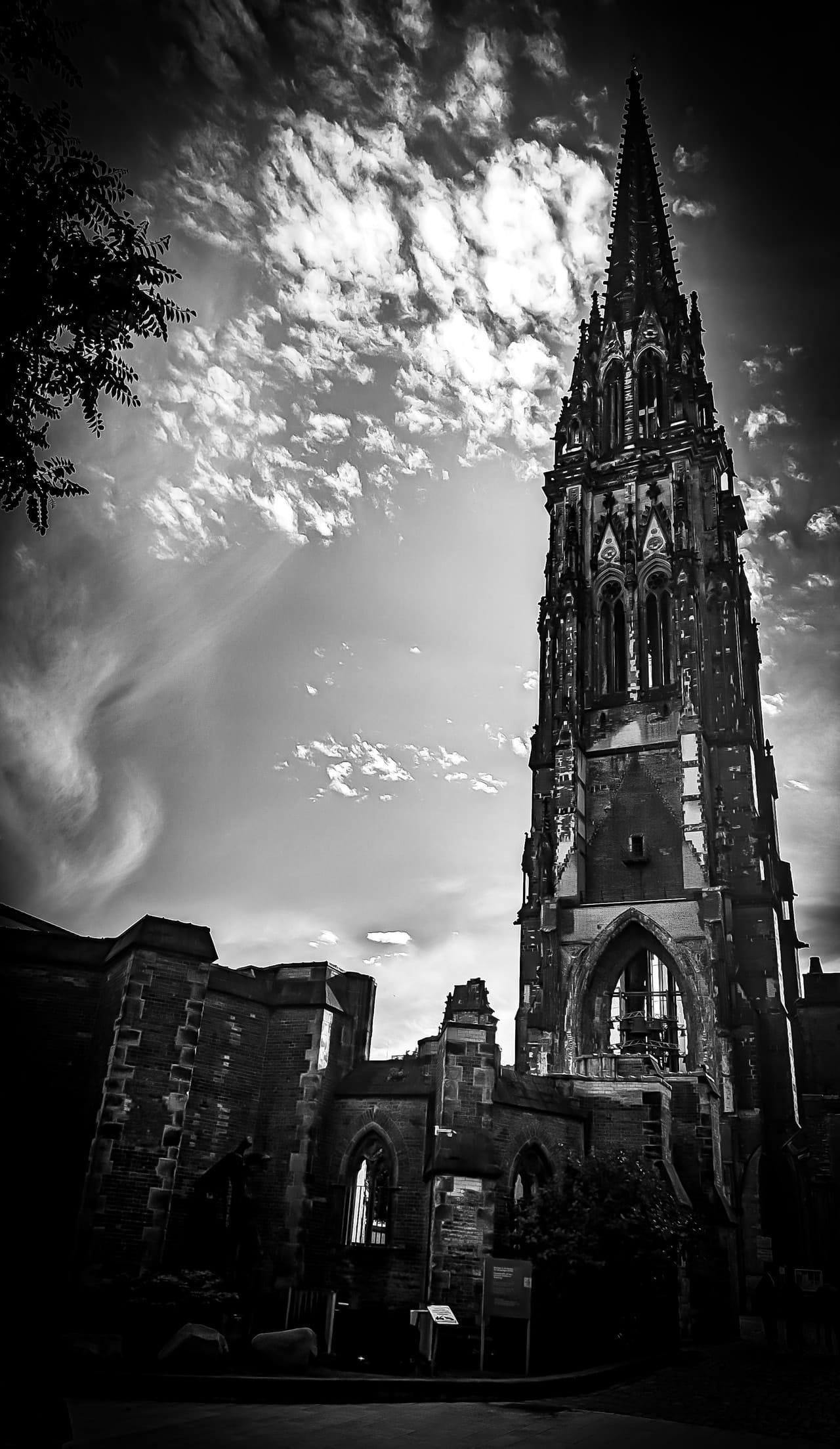 Black-and-white low-angle view of a Gothic church tower with an ornate spire rising on the right, attached to lower brick buildings with arched windows; cloud-filled sky overhead and tree branches silhouetted in the upper left.