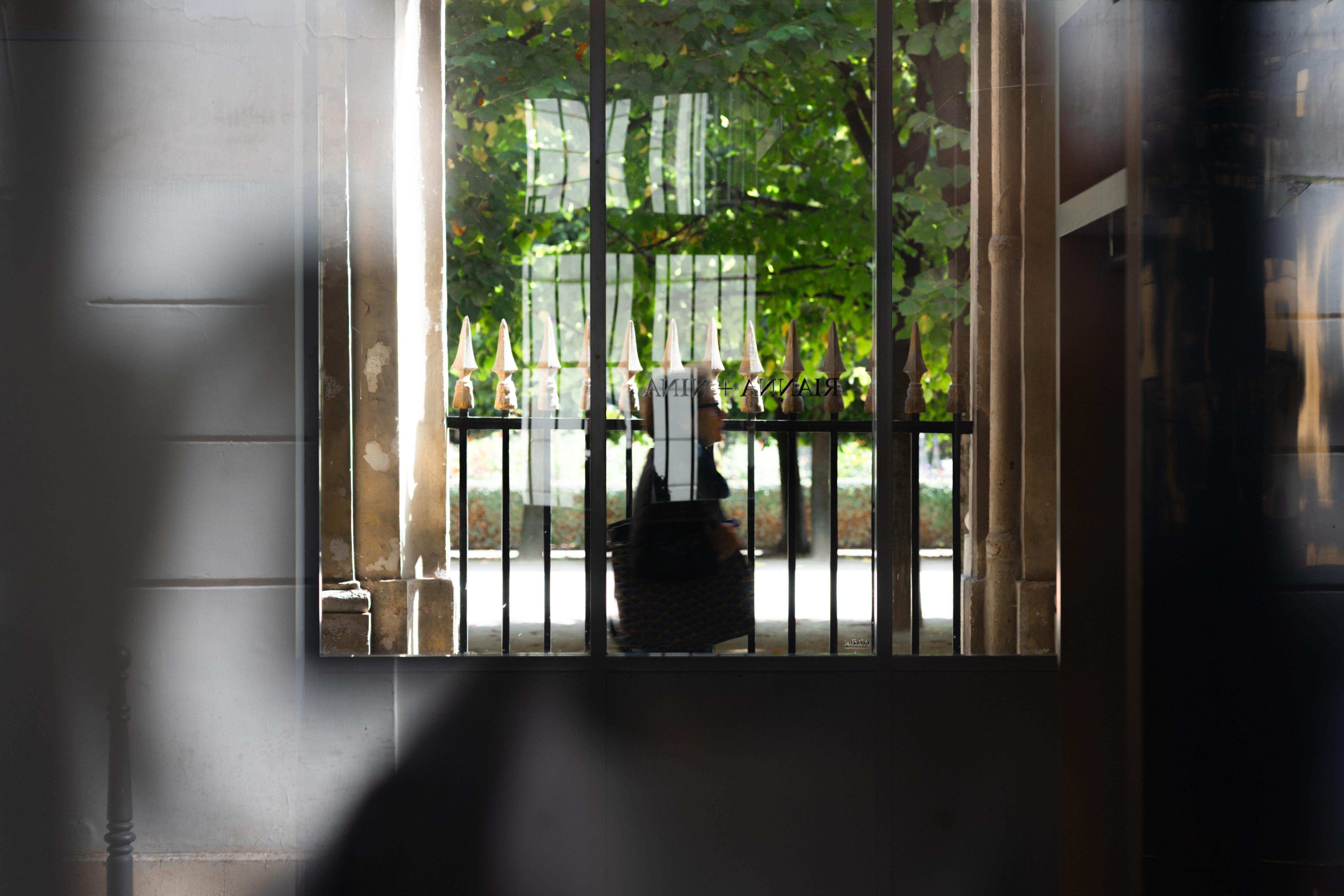 View of a window with a wrought iron fence outside, topped with pointed finials. Beyond the fence, a blurred silhouette of a person carrying a bag is visible. Lush green foliage and sunlight create a bright backdrop. The interior shadows from the window frame and walls add contrast to the scene.