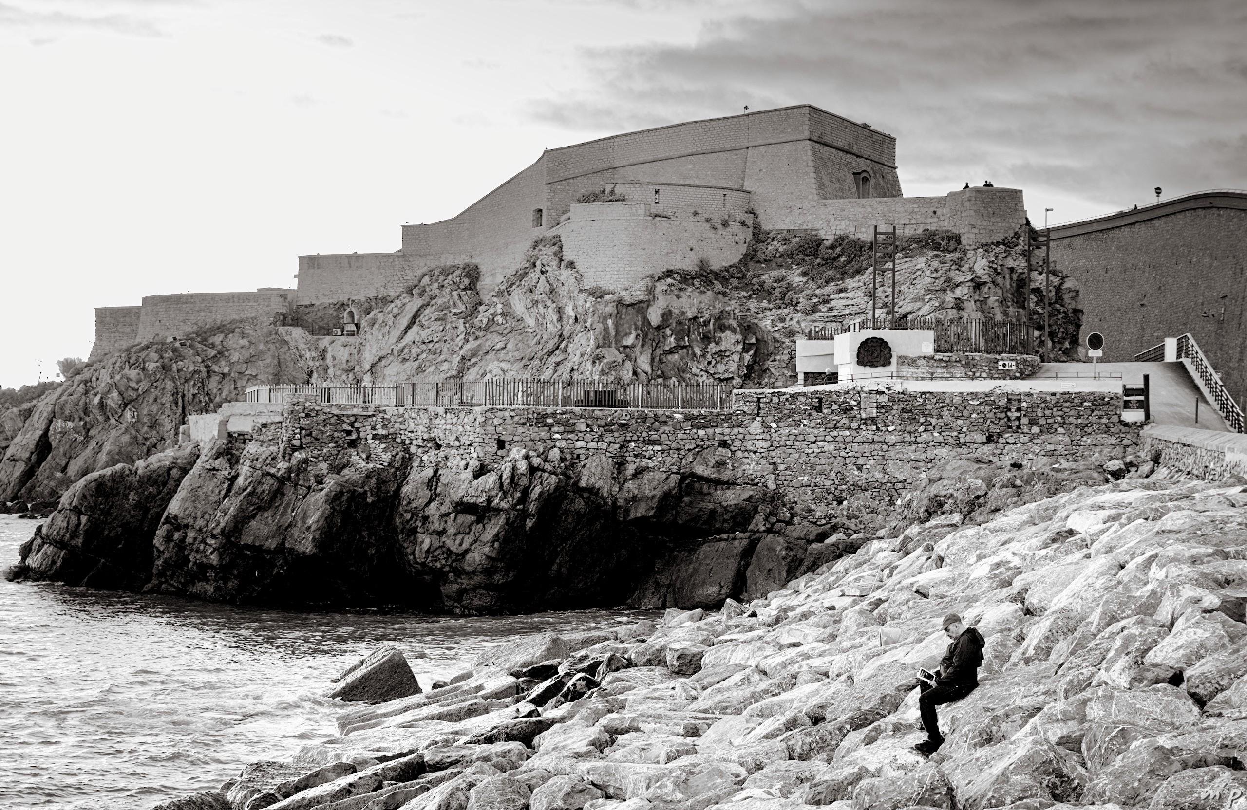 🇬🇧 Horizontal black and white photo of a man reading on a stone pier by the sea, under a castle built into the rock

///

🇫🇷 Photo noir et blanc horizontale d'un homme lisant sur une jetée de pierre en bord de mer, sous un château construit à même la roche