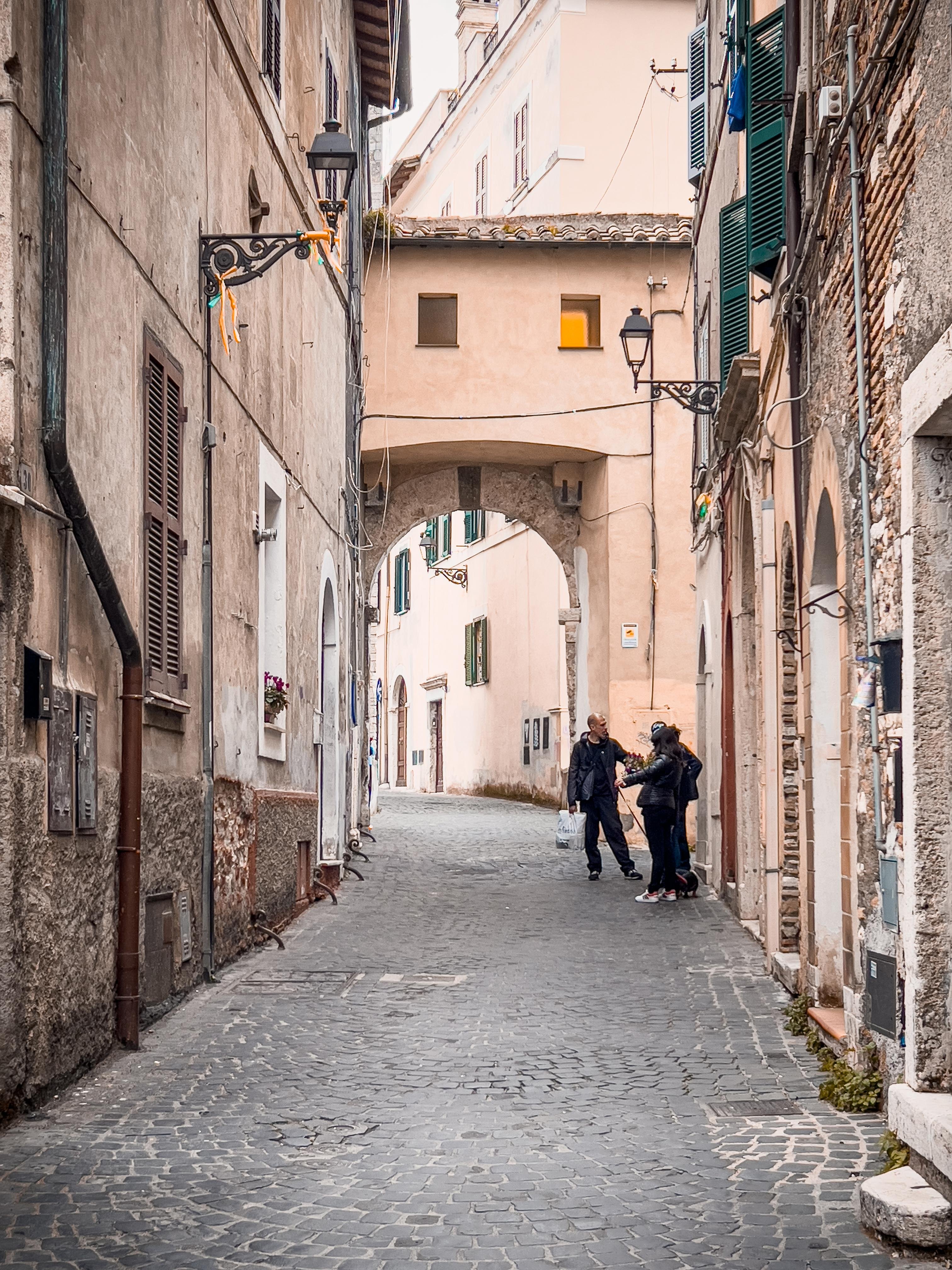 Stradina stretta e acciottolata in quella che sembra una città storica europea. Ci sono vecchi edifici su entrambi i lati, con facciate in pietra e intonaco, persiane in legno e lampioni in ferro decorati. Un arco collega due edifici sopra la strada. Alcune persone sono ferme e parlano sul lato destro della strada vicino a uno degli edifici. L'atmosfera appare calma e pittoresca.