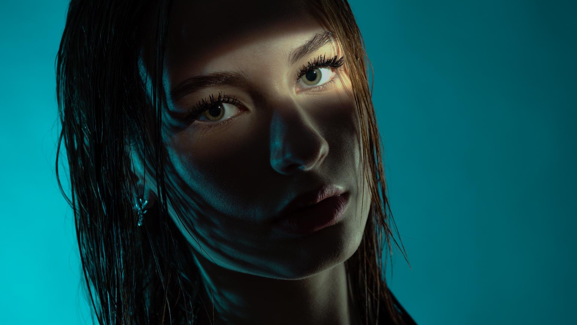A woman with wet hair stands against a teal background. Her face is partially illuminated by a narrow band of light, highlighting her eyes and lips. Shadows accentuate the contours of her face, creating a dramatic contrast. She wears an earring, visible on her left ear.