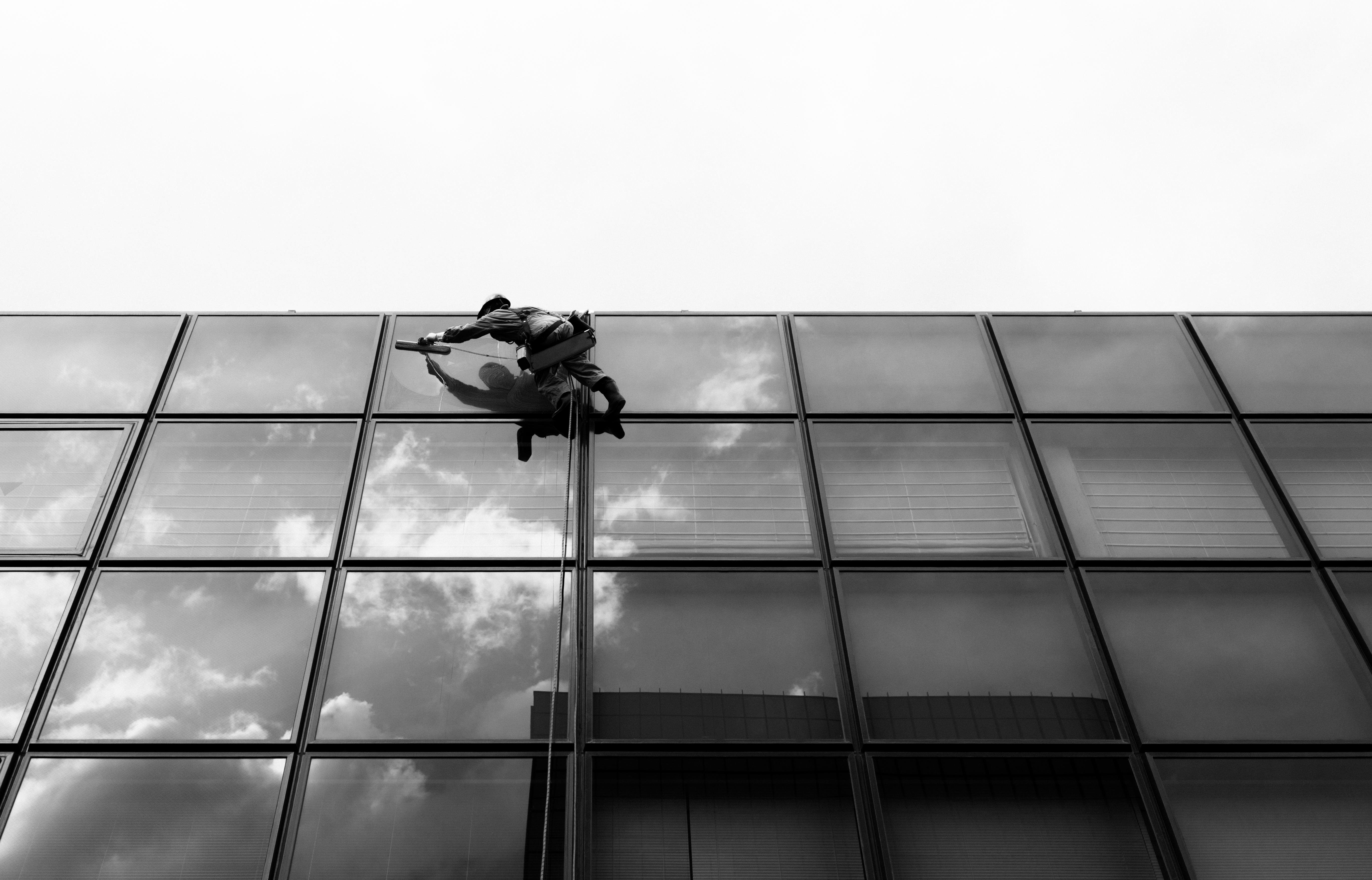 Person in climbing gear scaling a modern glass building facade, with reflections of clouds visible on the windows. The image is in black and white, emphasizing the geometric grid pattern of the windows.