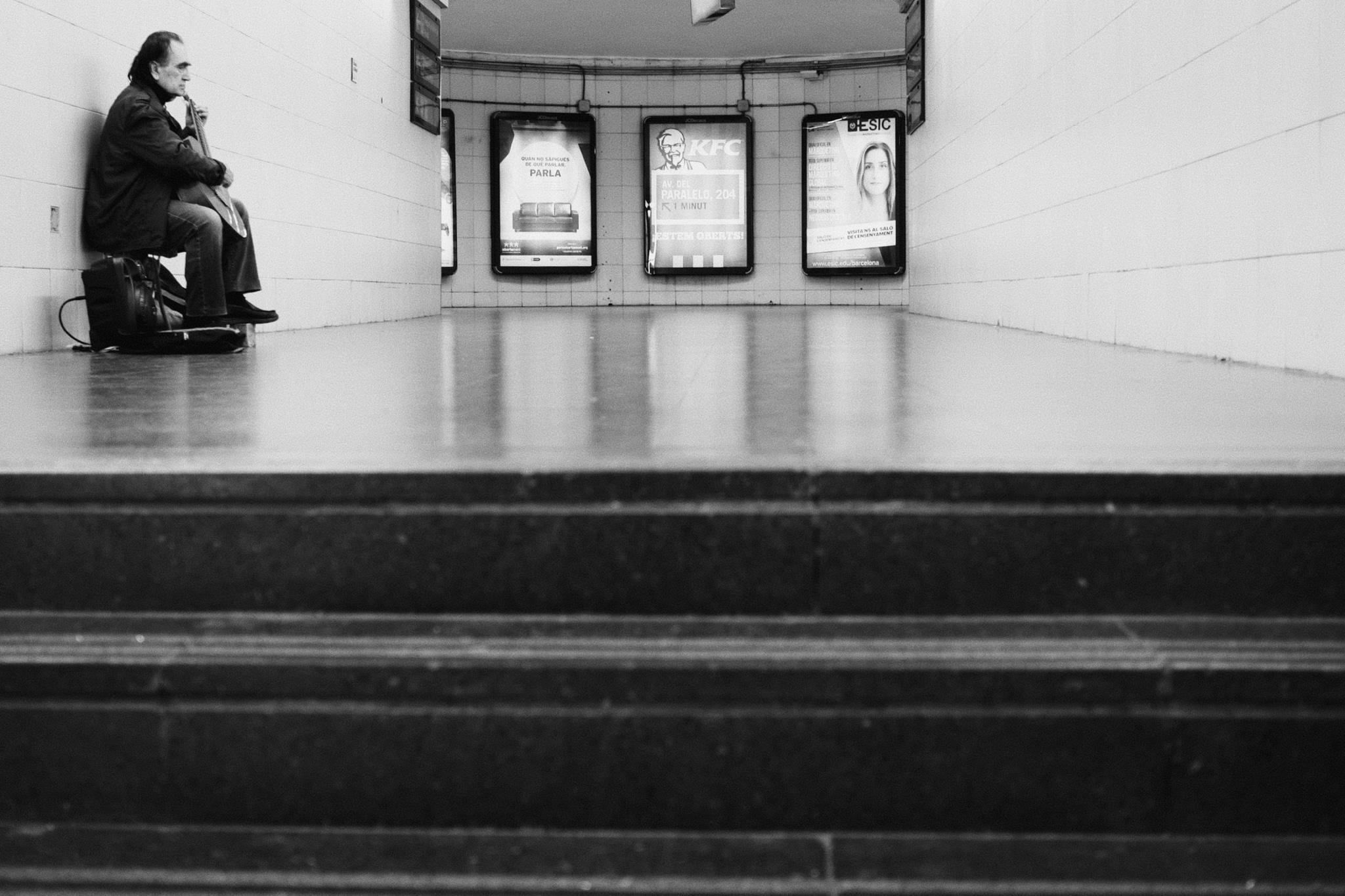 Schwarz-Weiss Foto. Mann sitzt in einen Durchgang auf der linken Seite und spielt mit einer Gitarre, am Ende des Durchgangs sind drei beleuchteten Werbeplakaten an der Rückwand. Im Vordergrund befindet sich breiten Treppe.