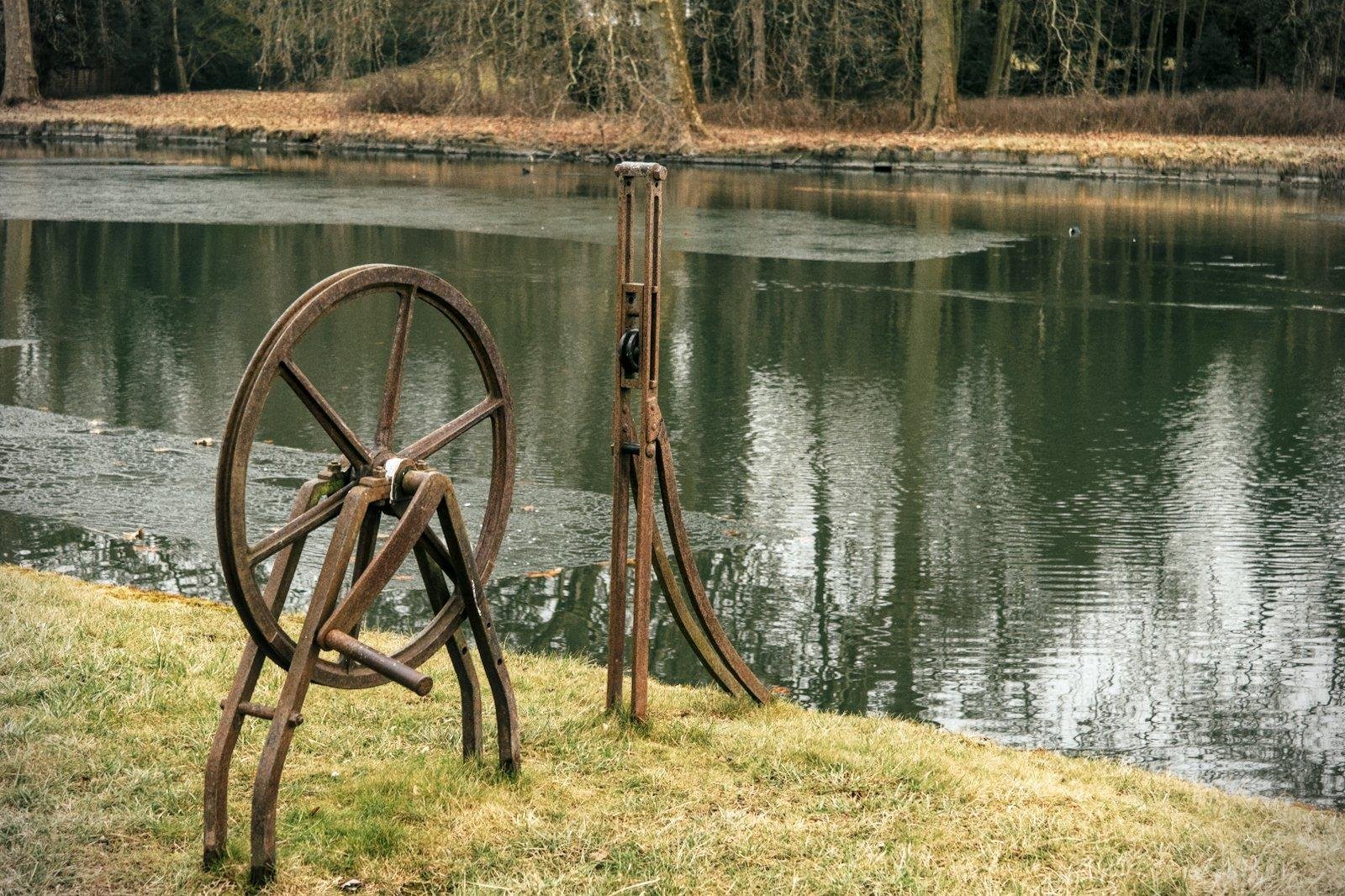 Photo format paysage montrant la rive d’un cours d’eau, avec de l’herbe jaunie, une roue en métal rouillée et un dispositif tout aussi rouillé qui pourrait être une poulie, dans le coin bas gauche. L’eau est verte et pleine de vaguelettes. L’arrière plan montre une forêt d’automne