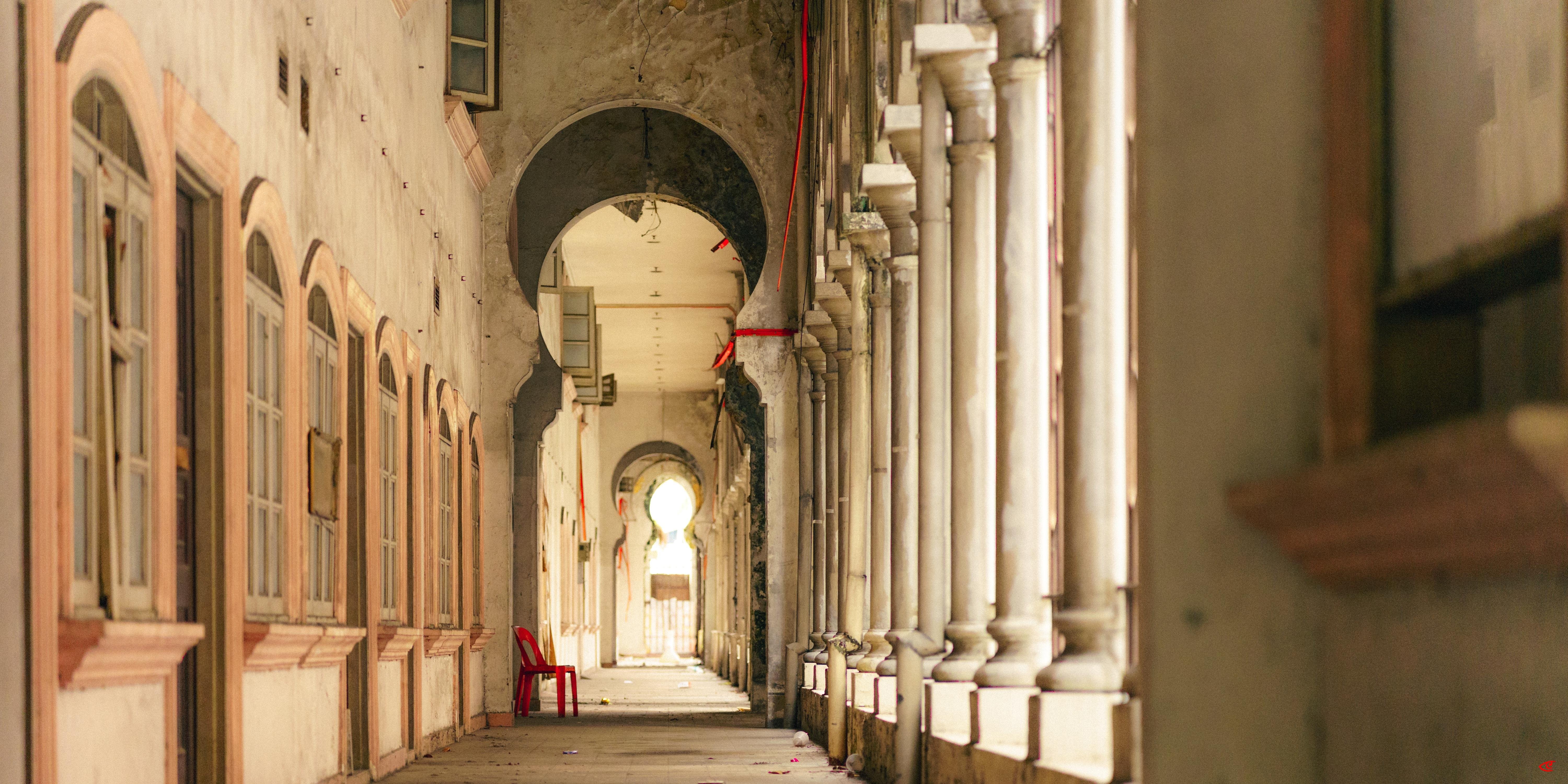 Long arched corridor with stone columns on the right and shuttered windows on the left, leading to a bright archway in the distance; a single red chair sits along the left side near the middle.