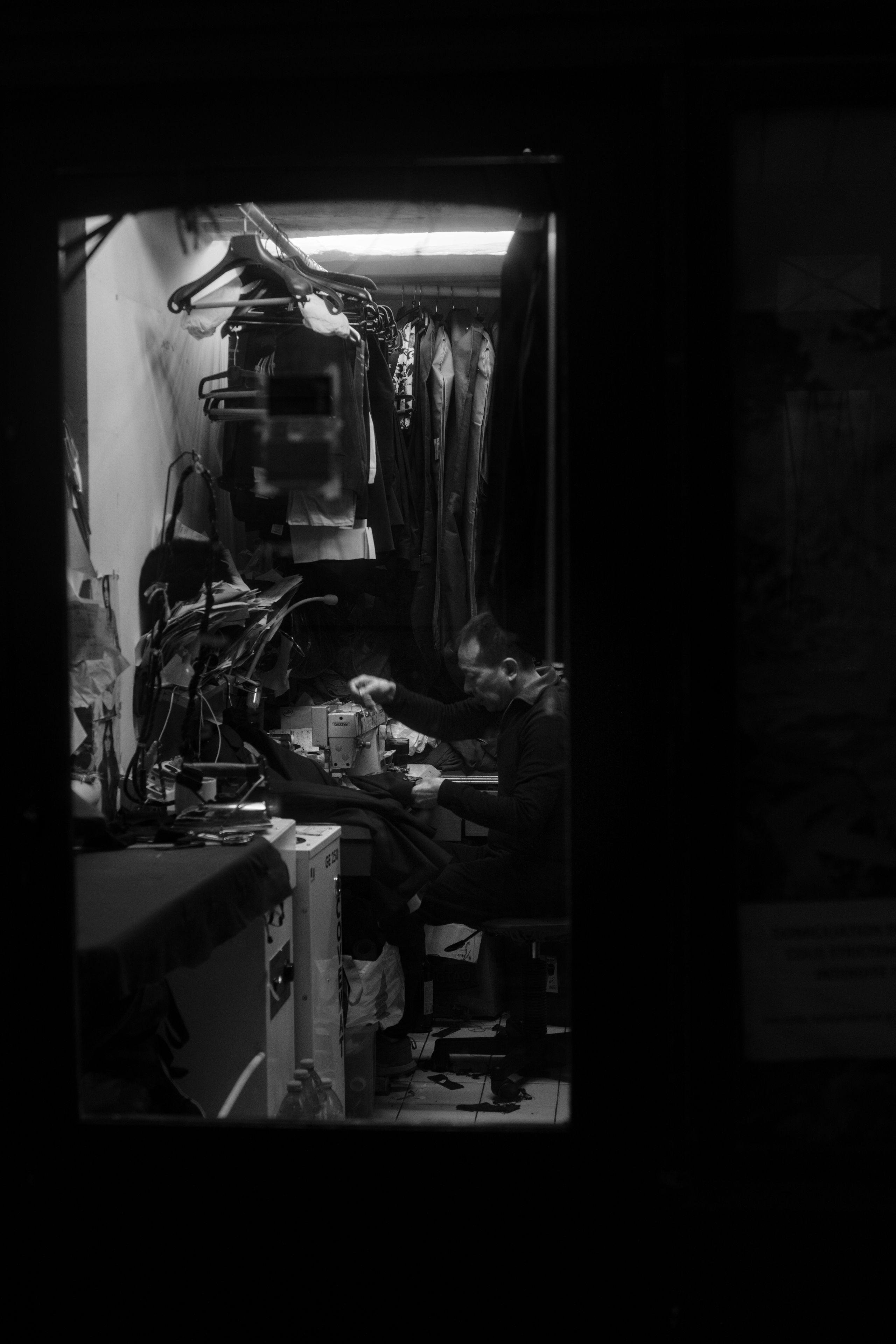Photo verticale en noir et blanc, de nuit. À travers la vitre d’une porte de boutique, un atelier de couture, des vestes accrochées en hauteur, un néon qui éclaire un homme travaillant devant une machine à coudre