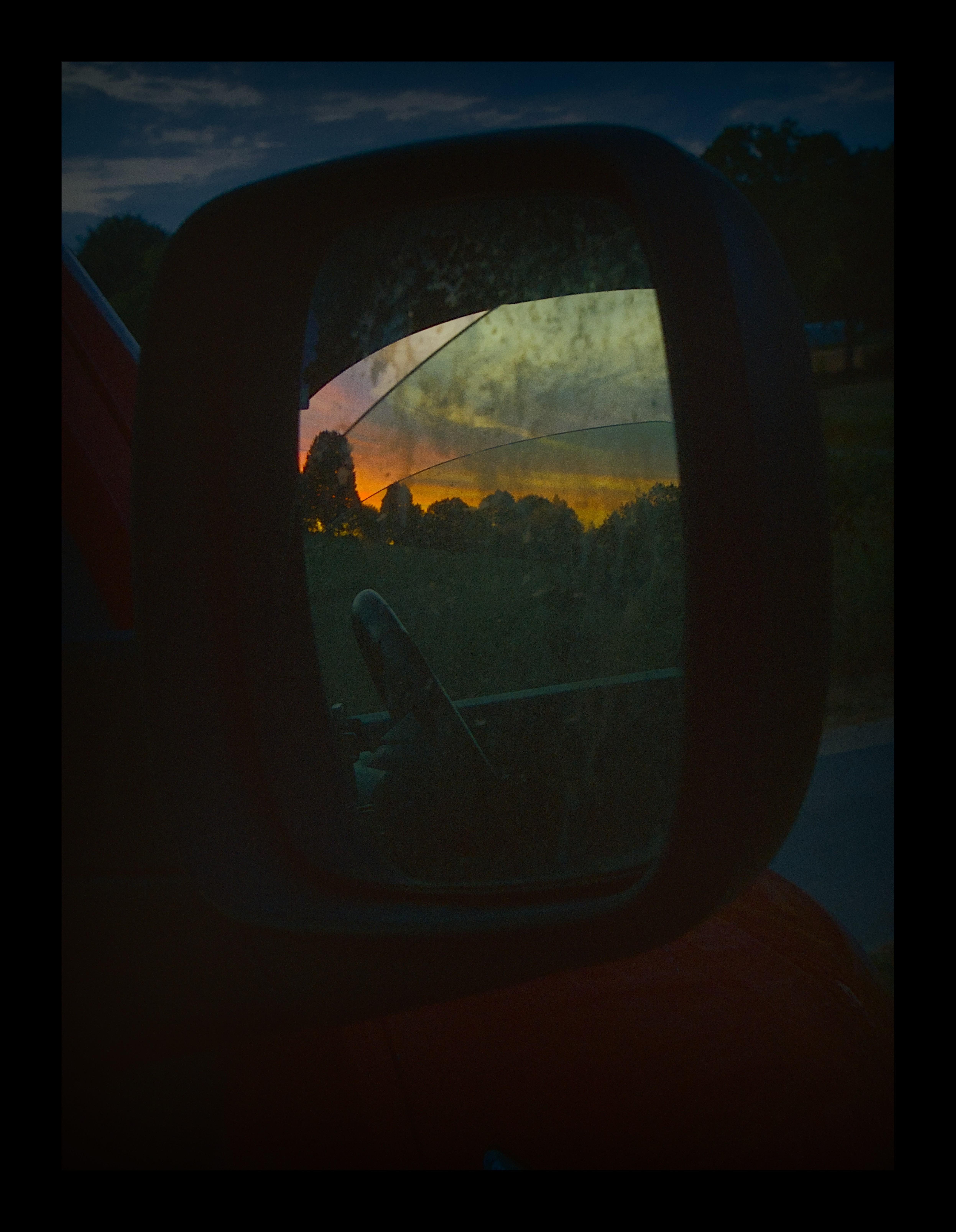 Car side-view mirror reflecting a sunset sky with orange and blue clouds above a dark tree line, with the mirror frame and part of the car door in shadow and streaks of dirt visible on the glass.