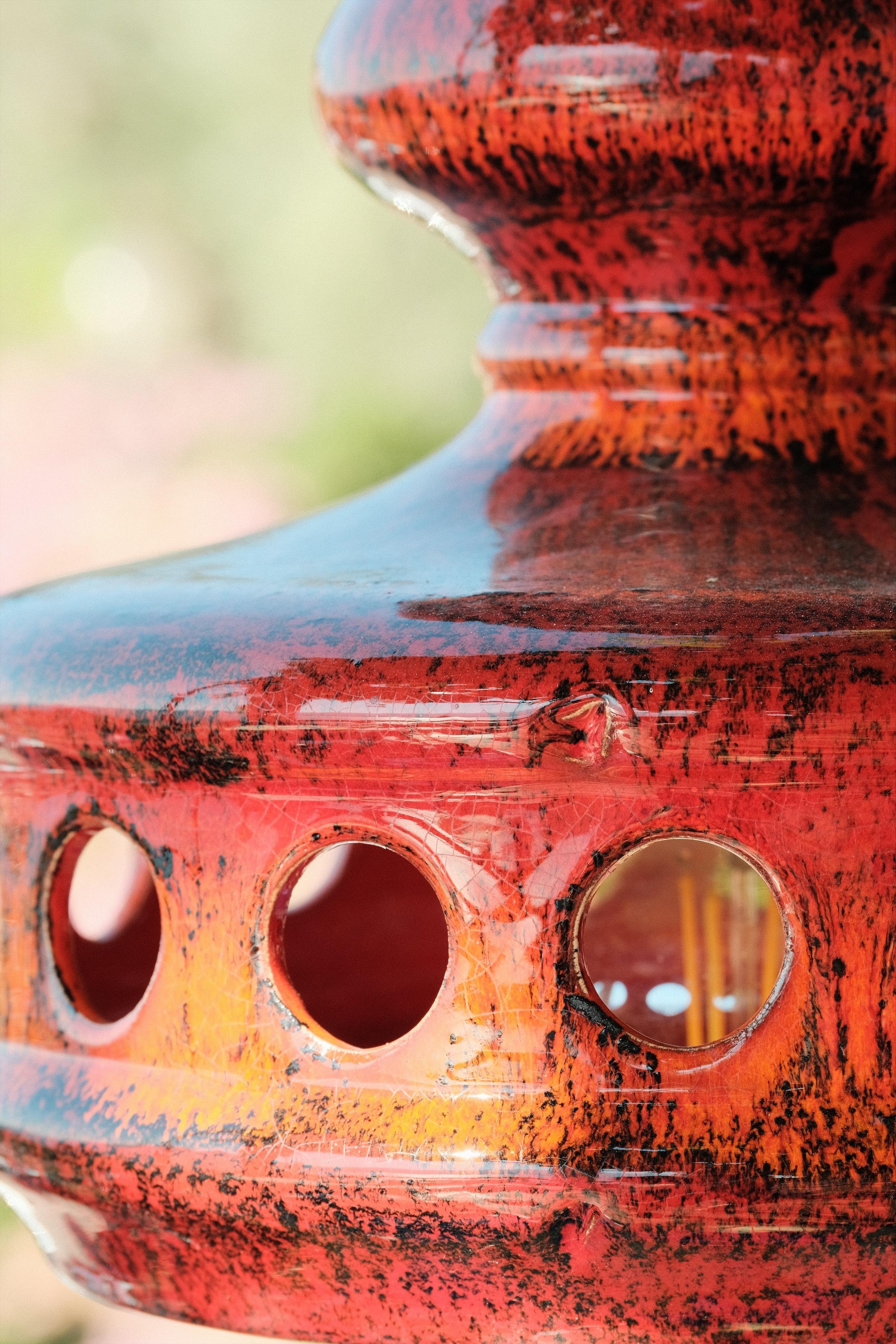 Detail Photo of a red/orange ceramic lamp with black sprinkles. Through round holes you can see the lightbulb. The lamp would have a bell shape, hanging from ceiling of a terrace. Here you can only see a part of it.